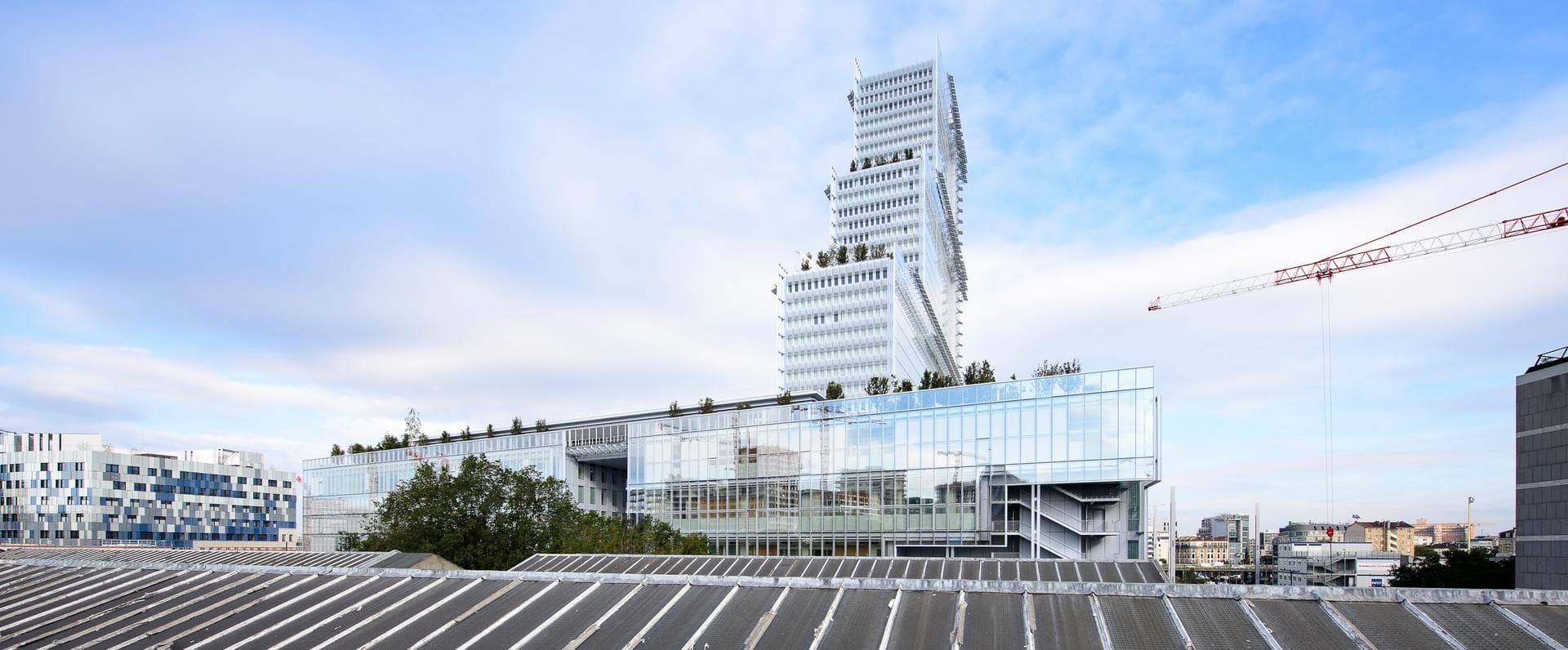 Un bâtiment moderne blanc et vitré avec une base large et une tour étagée ornée de toits végétalisés se dresse sous un ciel nuageux. Au premier plan, un toit gris à bandes ; une grue de construction rouge et blanche est visible à droite.
