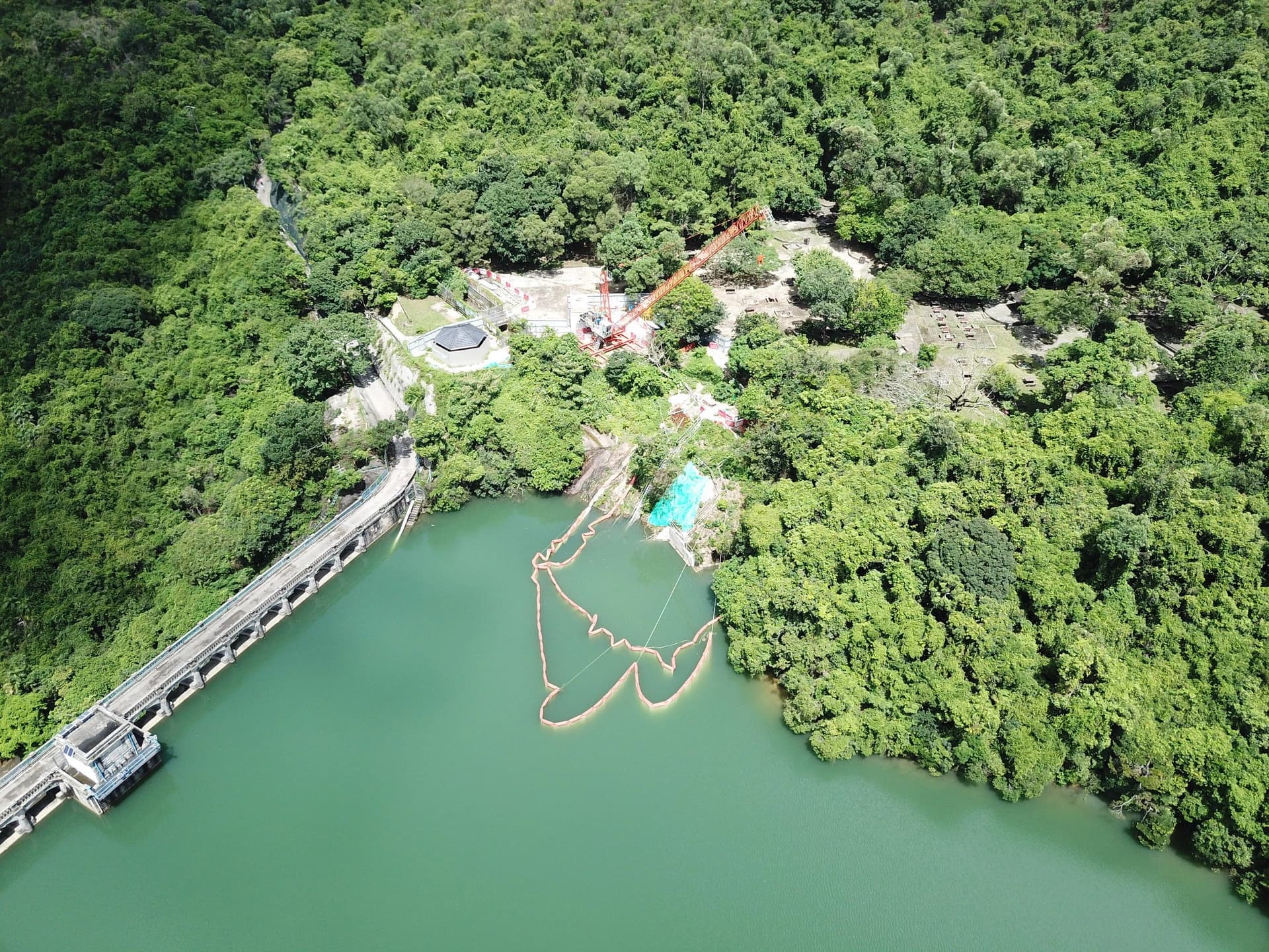 Vue aérienne d'un réservoir vert émeraude avec un barrage en béton incurvé à gauche, bordé par des forêts luxuriantes. Sur la rive droite, un chantier avec une grue orange et des barrières flottantes dans l'eau est visible.