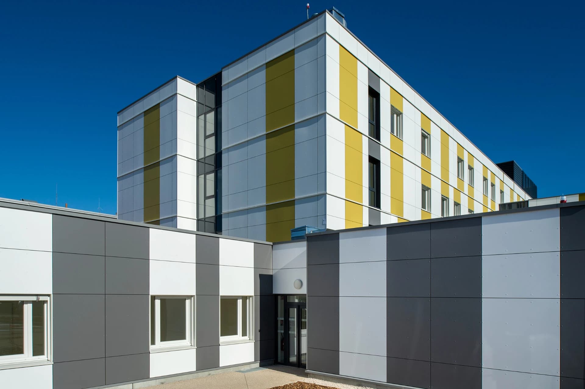 Un bâtiment moderne à plusieurs étages présente une façade avec des rayures verticales alternées blanches, gris clair et chartreuse, partiellement masqué par une structure inférieure aux panneaux horizontaux gris foncé, gris clair et blancs. Le tout est sous un ciel bleu clair éclatant.