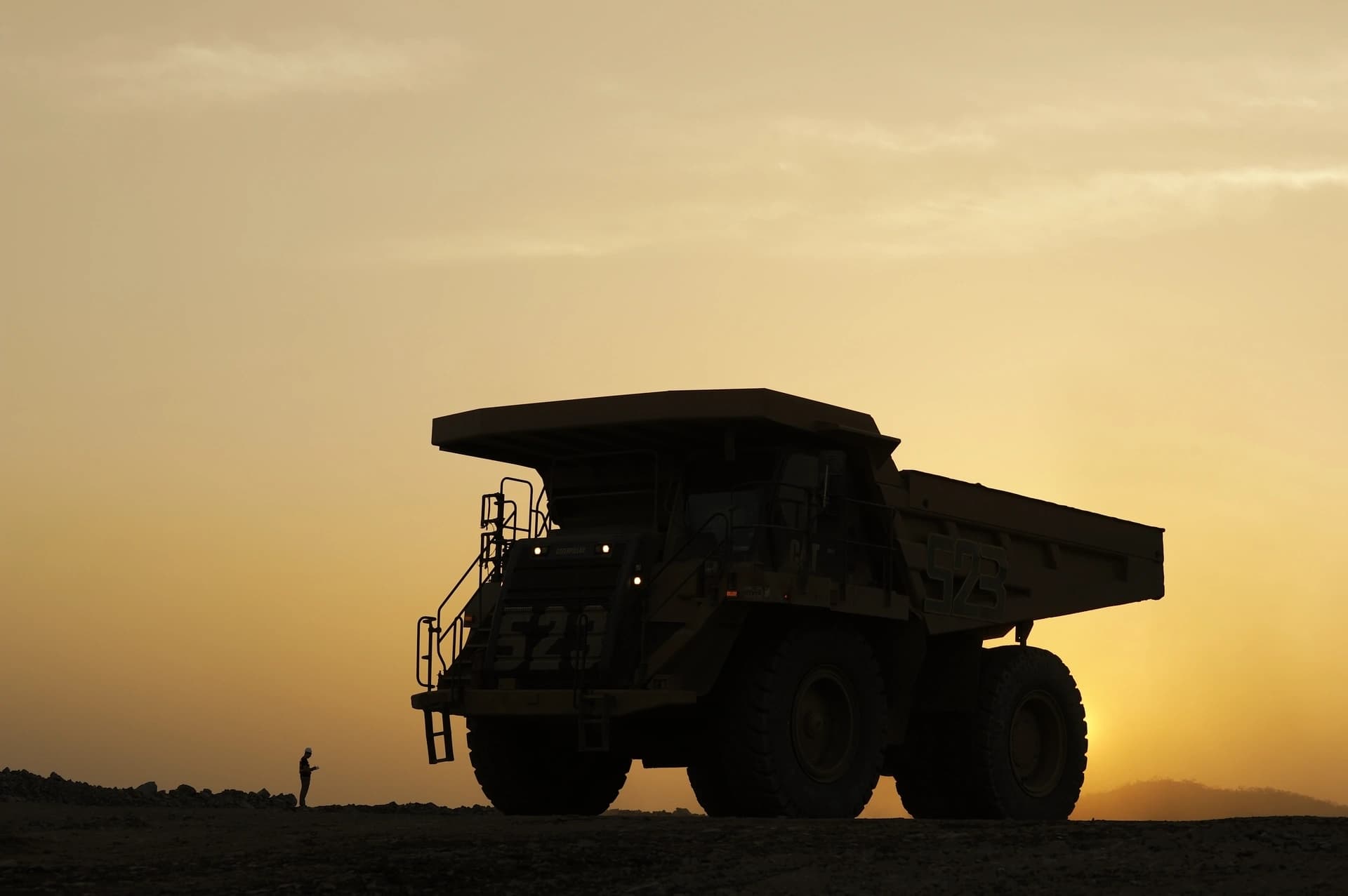 Silhouette d'un immense camion minier stationné à côté d'une personne isolée sur un terrain accidenté, baigné par les teintes chaudes d'un ciel orange et jaune au crépuscule.