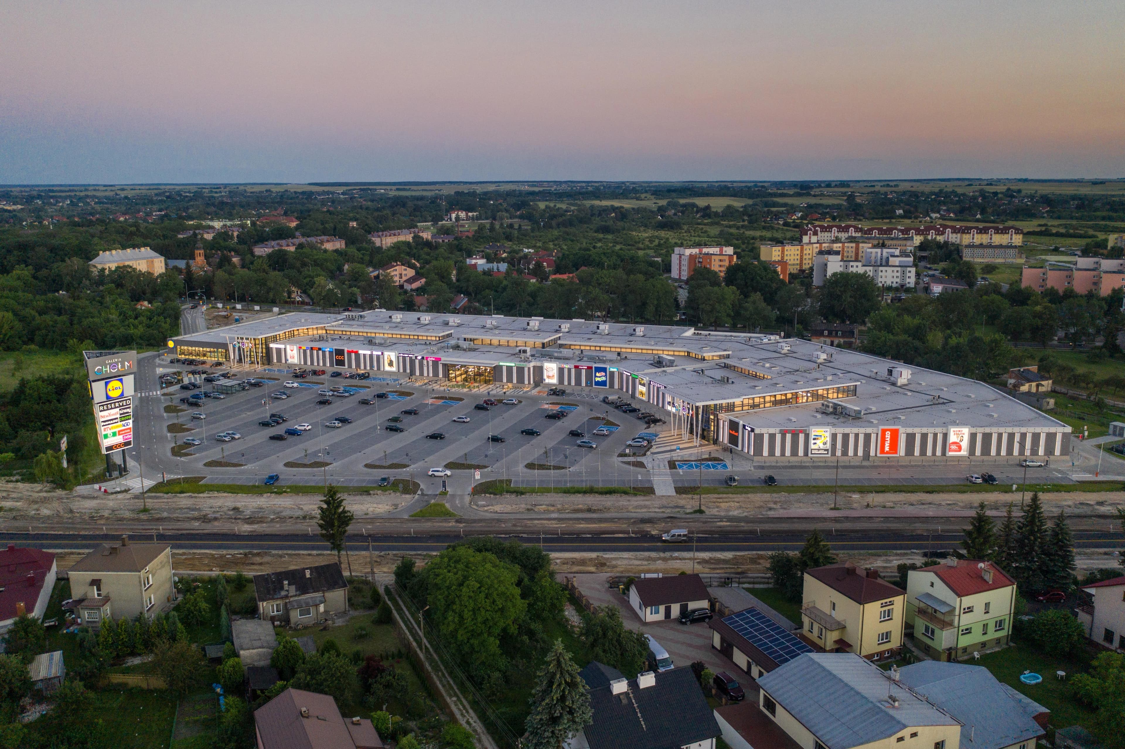 Vue aérienne d'un vaste centre commercial moderne, la Galeria Chełm, avec un grand parking rempli de voitures, au crépuscule. Le bâtiment aux enseignes lumineuses est entouré de zones résidentielles et d'arbres, sous un ciel dégradé de rose et de bleu.