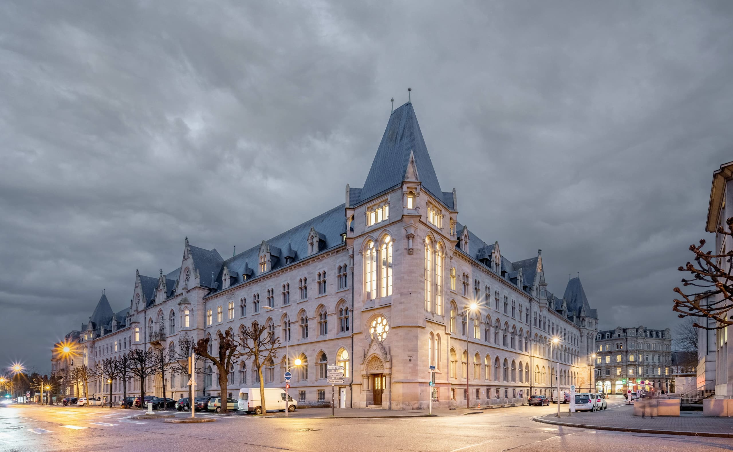 Grand bâtiment en pierre grise avec de hauts toits d'ardoise, ses fenêtres éclairées d'une lumière dorée au crépuscule, domine une rue urbaine calme avec des voitures et des arbres dénudés sous un ciel nuageux.
