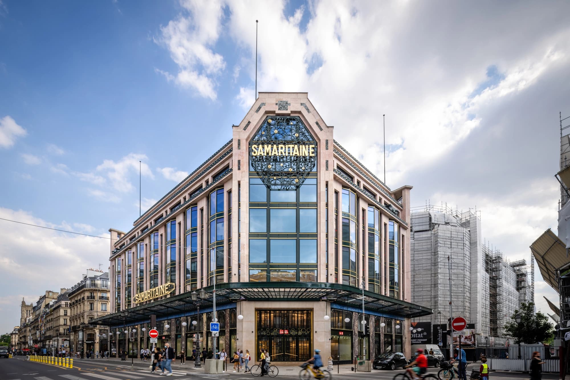 La façade élégante du grand magasin La Samaritaine à Paris, un bâtiment beige et vert avec de grandes fenêtres et des enseignes dorées, sous un ciel partiellement nuageux avec des passants dans la rue.