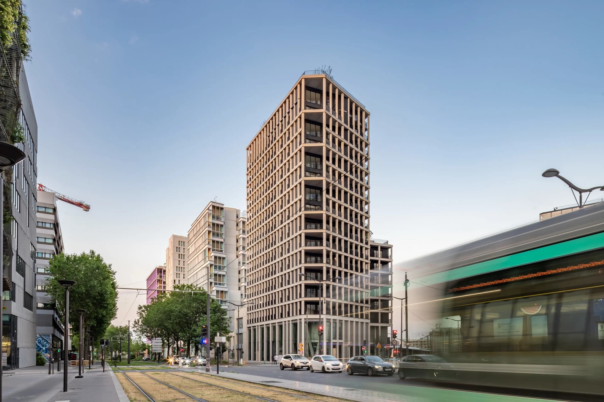 Scène urbaine moderne avec un grand immeuble beige à façade quadrillée au centre, bordé d'autres bâtiments contemporains sous un ciel bleu clair. Un tram vert et gris foncé flou passe rapidement à droite, tandis que des voitures circulent sur la route à côté de rails de tramway et d'arbres.
