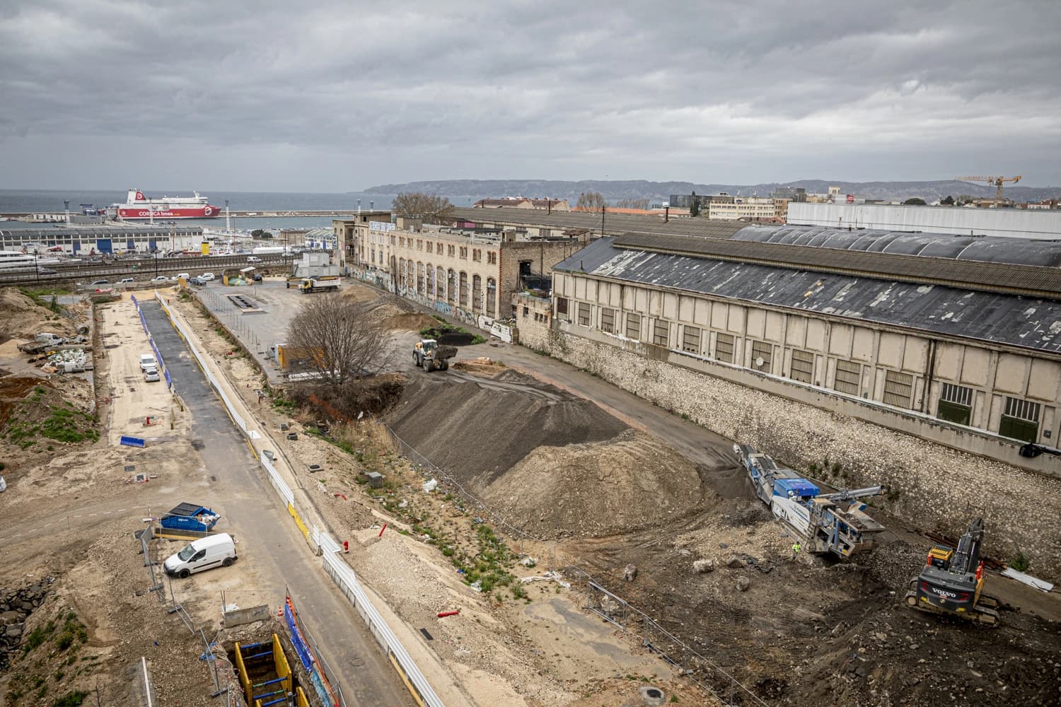 Vue aérienne d'un vaste chantier de construction urbain avec des engins de terrassement et des monticules de terre, des bâtiments industriels vieillissants, et un ferry rouge amarré dans un port sous un ciel nuageux.