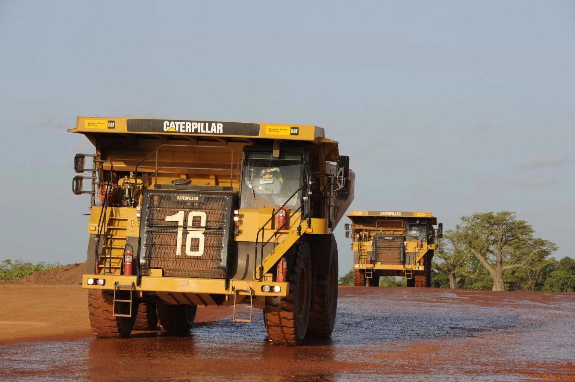 Deux énormes camions miniers jaunes Caterpillar avancent sur une route de terre rouge et humide, le premier portant le numéro 18 avec un conducteur visible. Un second camion suit au loin sous un ciel clair, avec quelques arbres éparses à l'horizon.
