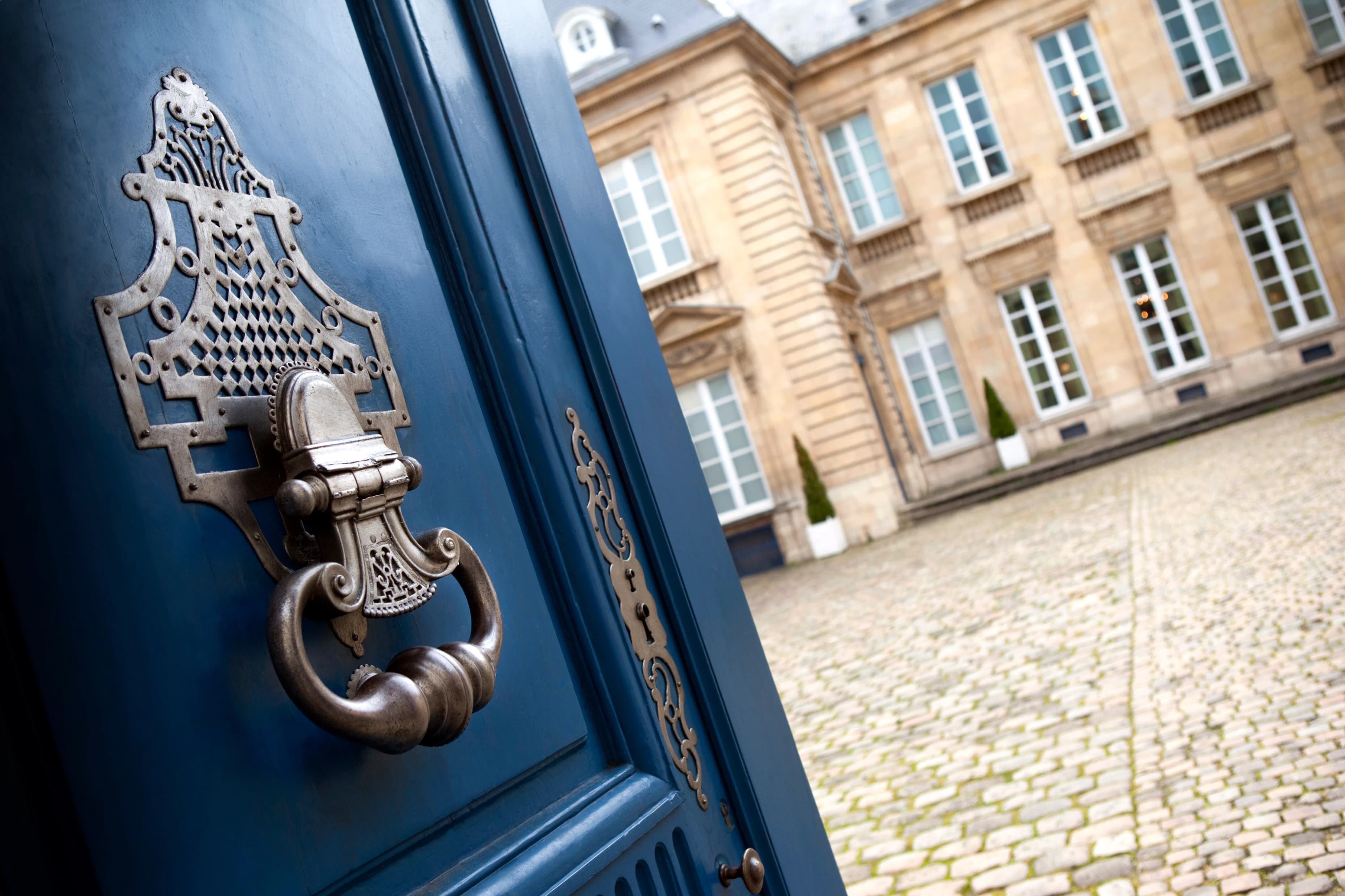 Une porte sculptée d'un bleu profond, ornée d'un grand heurtoir en métal argenté et d'un médaillon de serrure, s'entrouvre sur une cour pavée et un élégant bâtiment en pierre beige avec de nombreuses fenêtres.