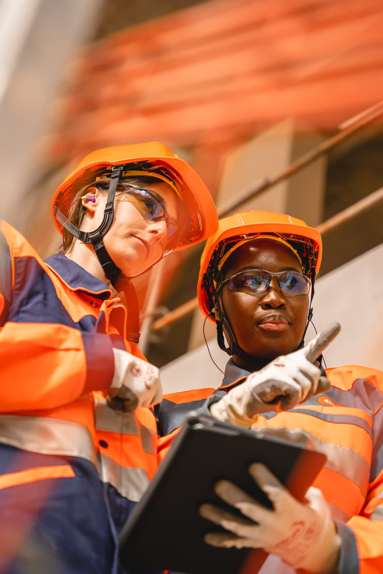 Deux professionnelles portant des casques orange, des lunettes de sécurité et des uniformes de travail orange et bleu sont sur un chantier, l'une tenant une tablette et pointant vers quelque chose tandis que l'autre observe.