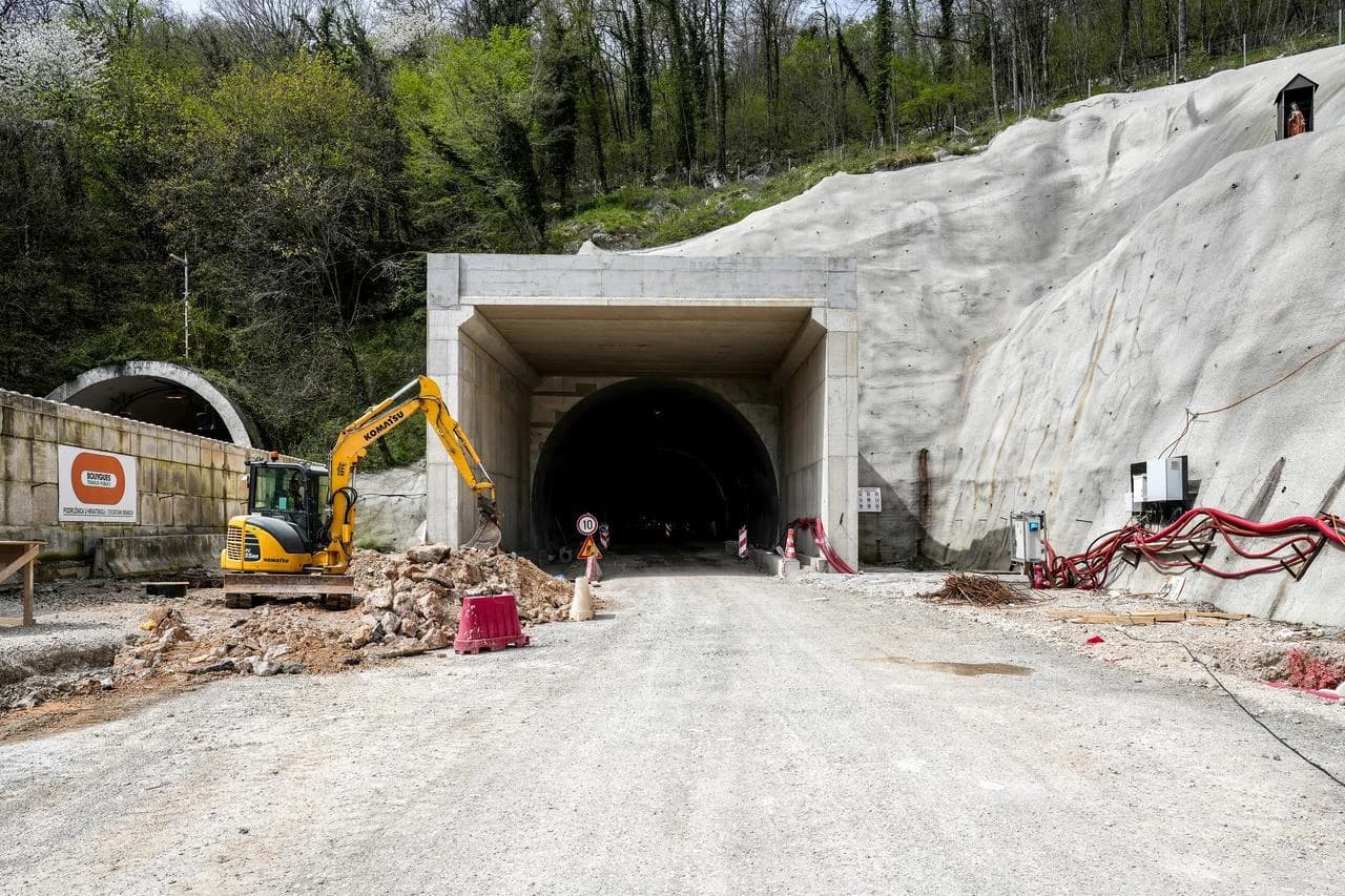 Vue d'un chantier de construction avec l'entrée sombre d'un grand tunnel en béton gris au centre, encadrée par un mur de soutènement similaire d'où pendent des câbles rouges épais. Un excavateur jaune Komatsu est visible à gauche déblayant des roches, tandis que l'arrière-plan montre des collines verdoyantes et une entrée de tunnel secondaire.