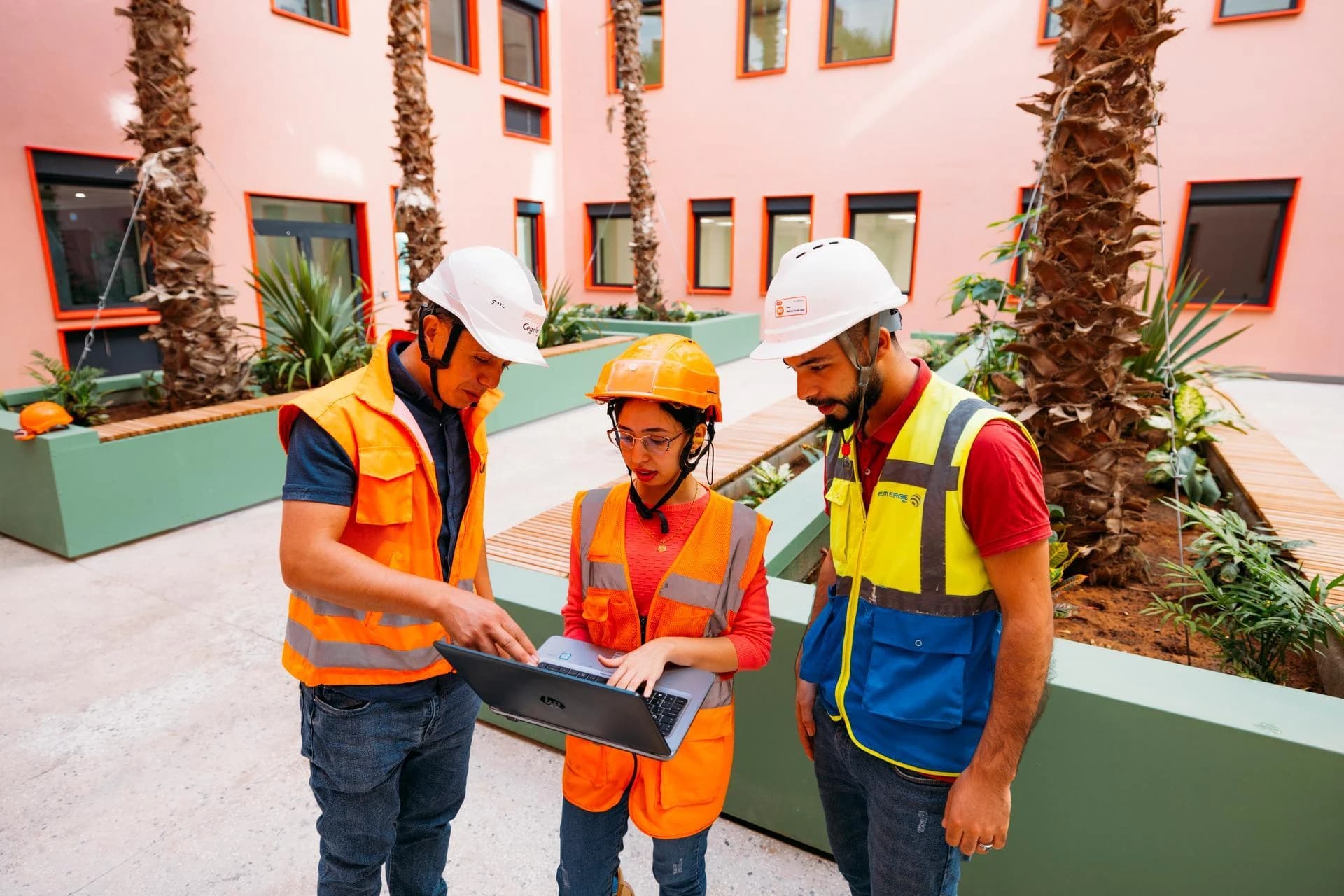 Trois professionnels en casques et gilets de sécurité orange et jaune examinent un ordinateur portable dans la cour d'un bâtiment rose moderne, entourés de verdure et de palmiers.