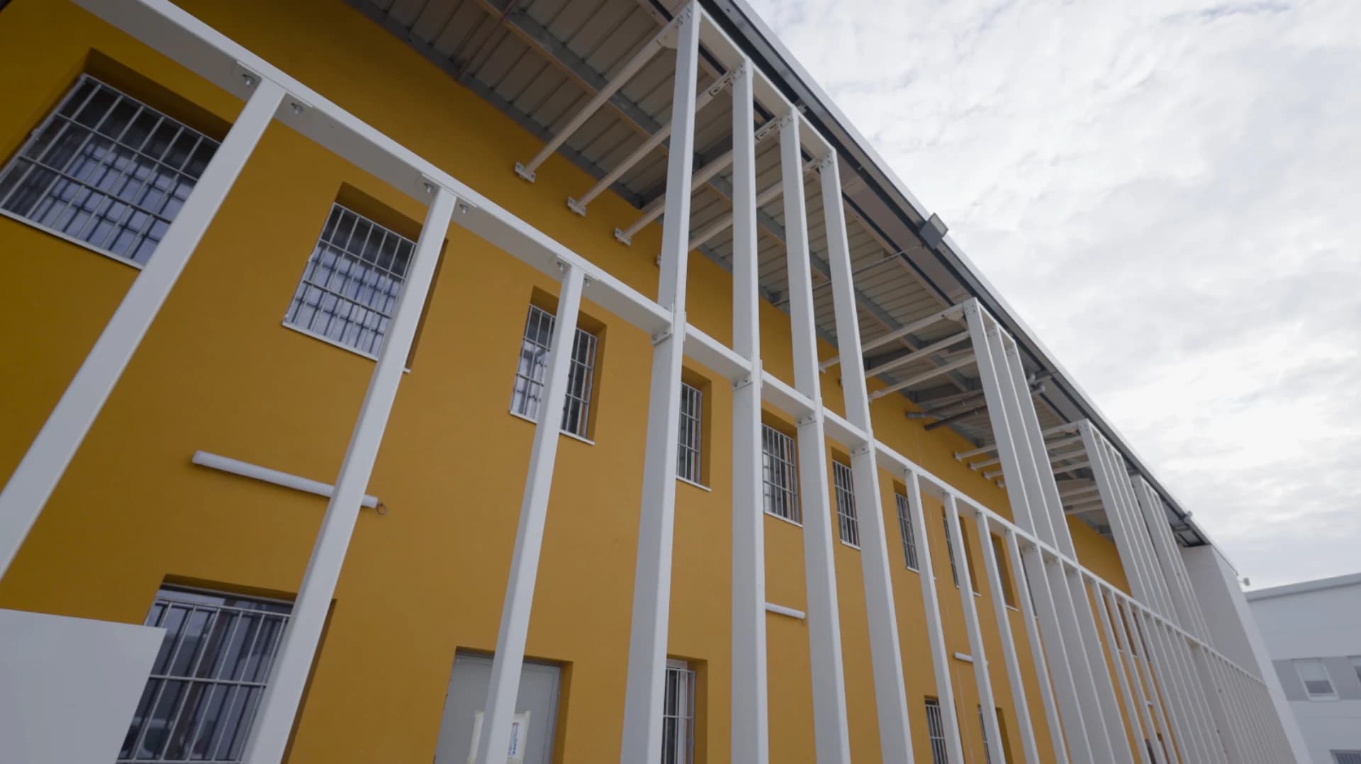 Façade moderne d'un bâtiment jaune vif vue d'en bas, présentant plusieurs fenêtres grillagées et une imposante structure en métal blanc s'étendant verticalement sous un ciel nuageux.