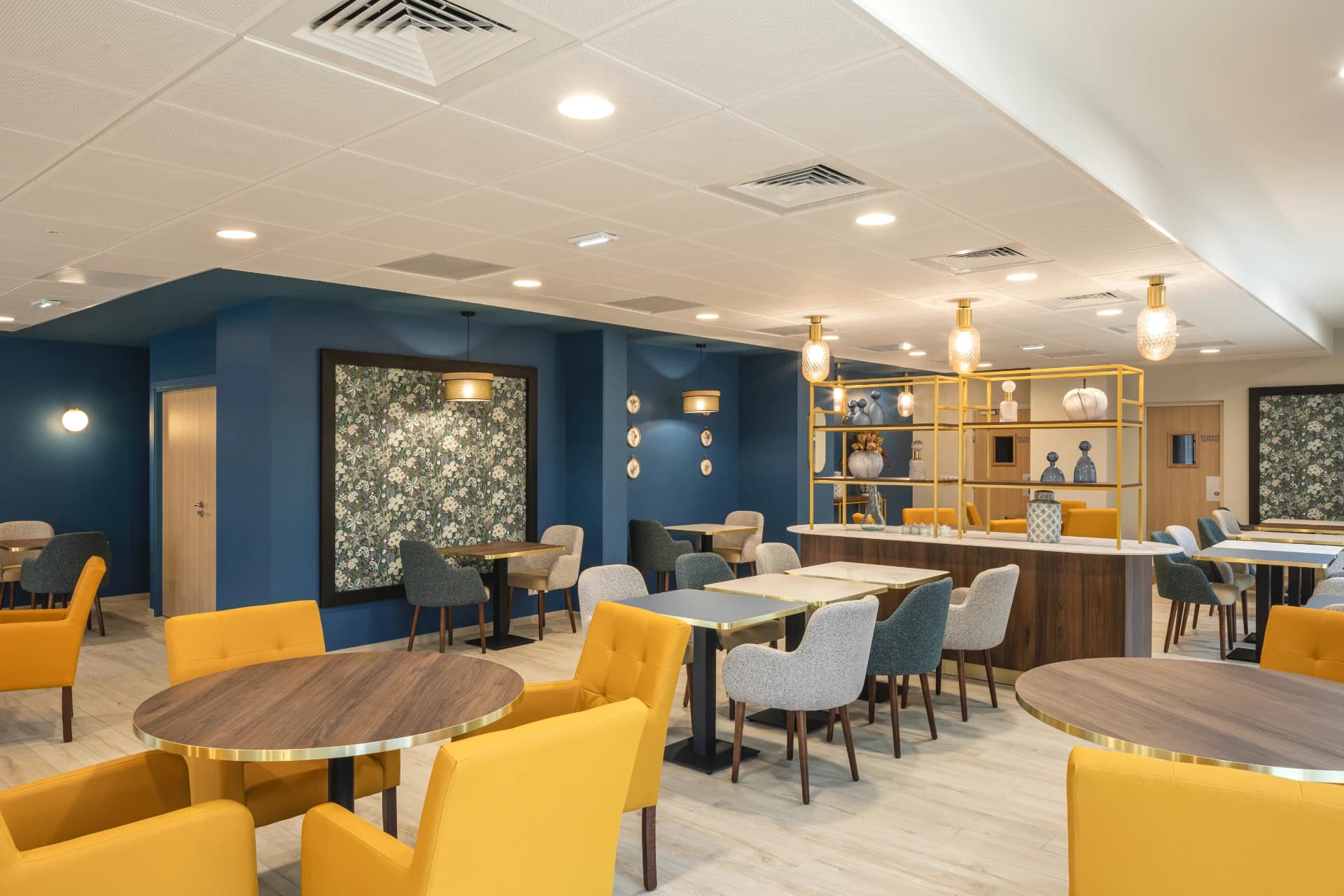 Un espace de restauration ou de salon moderne et lumineux, avec des murs bleu foncé, un grand tableau floral et un sol en bois clair. Il est aménagé avec diverses tables et un mélange de fauteuils jaune moutarde et de chaises en tissu gris.