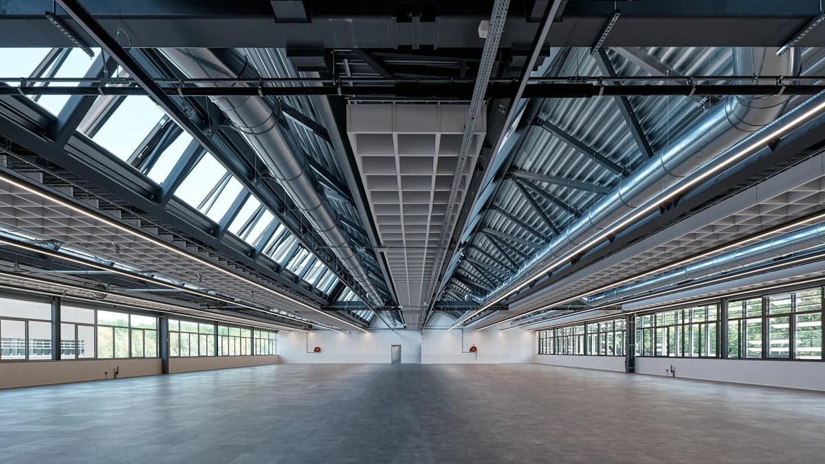 Vaste hall moderne et vide avec un haut plafond industriel exposé en noir et argent, éclairé par des puits de lumière et de longs luminaires LED. Les murs blancs sont percés de grandes fenêtres offrant une vue sur l'extérieur et le sol est en carrelage gris.