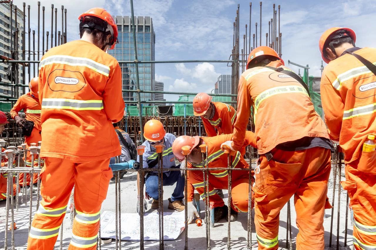 Des ouvriers du bâtiment en uniforme orange et casque de sécurité travaillent sur un chantier, certains examinant des plans au milieu d'armatures métalliques.