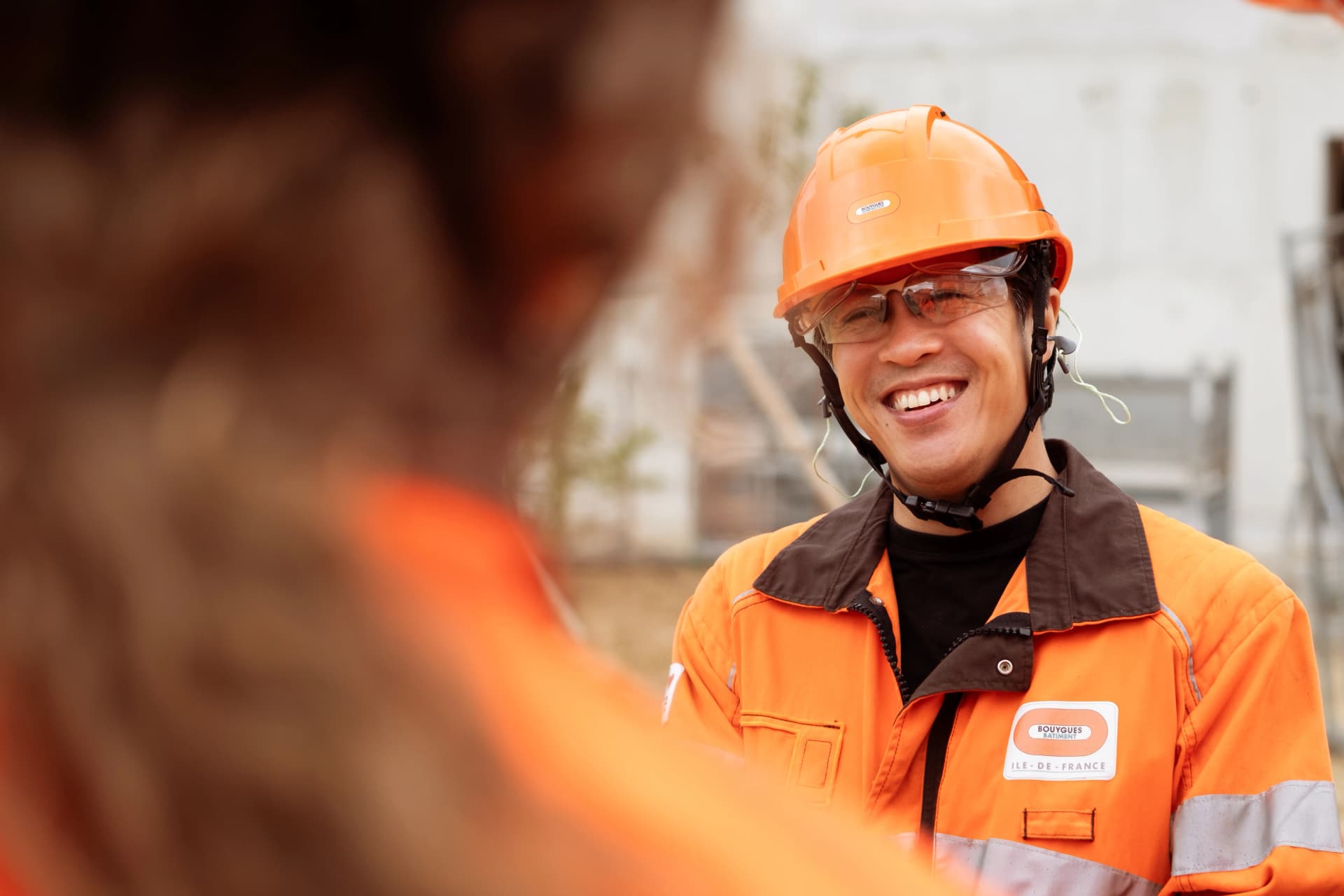 Un homme souriant, portant un casque de sécurité orange et une veste de chantier haute visibilité Bouygues Bâtiment Île-de-France, regarde vers une personne floue au premier plan.