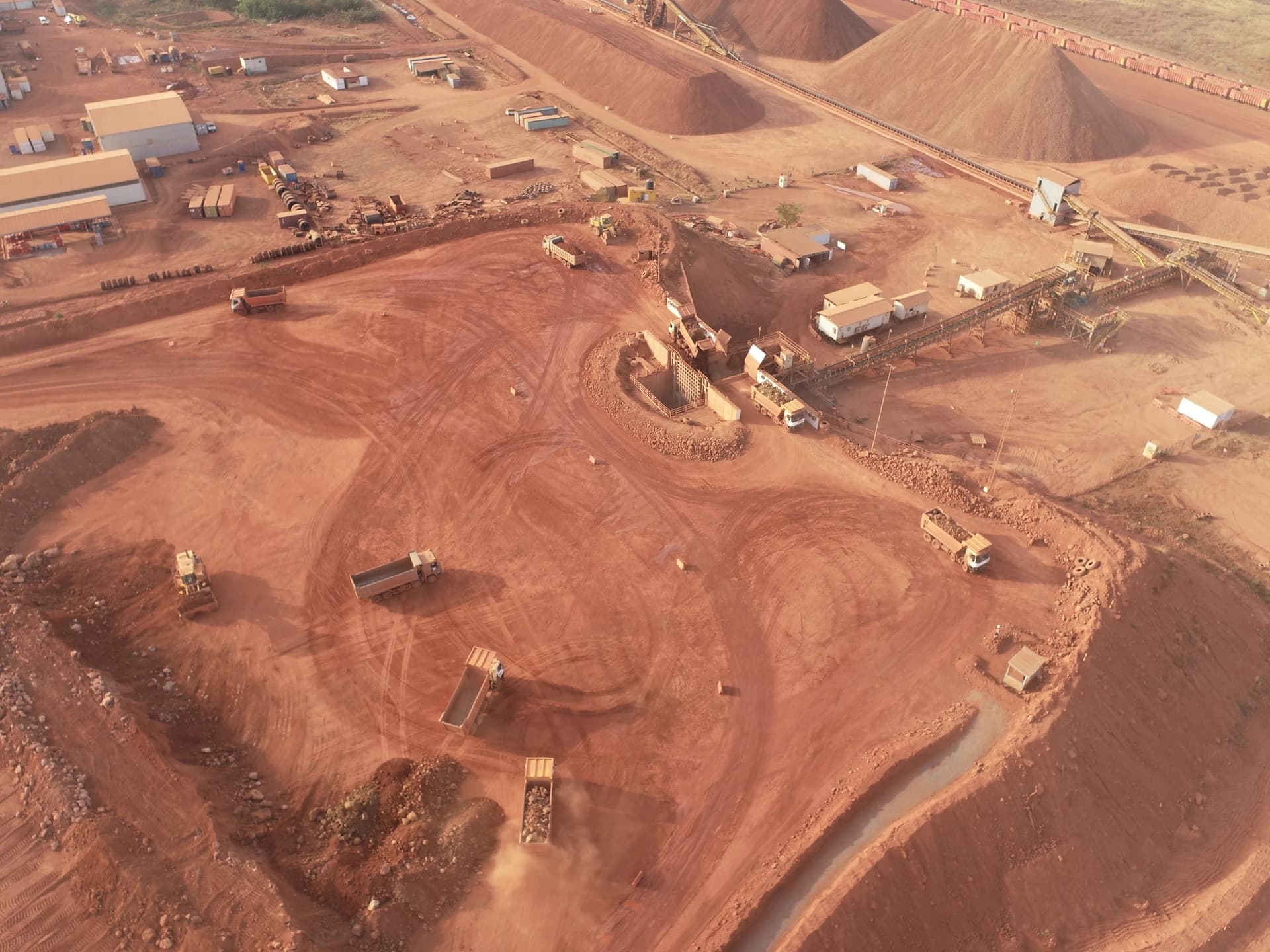 Vue aérienne d'une vaste mine à ciel ouvert de couleur rouille, montrant des camions-bennes actifs, des convoyeurs et d'énormes tas de minerai sur un paysage terreux et rouge.