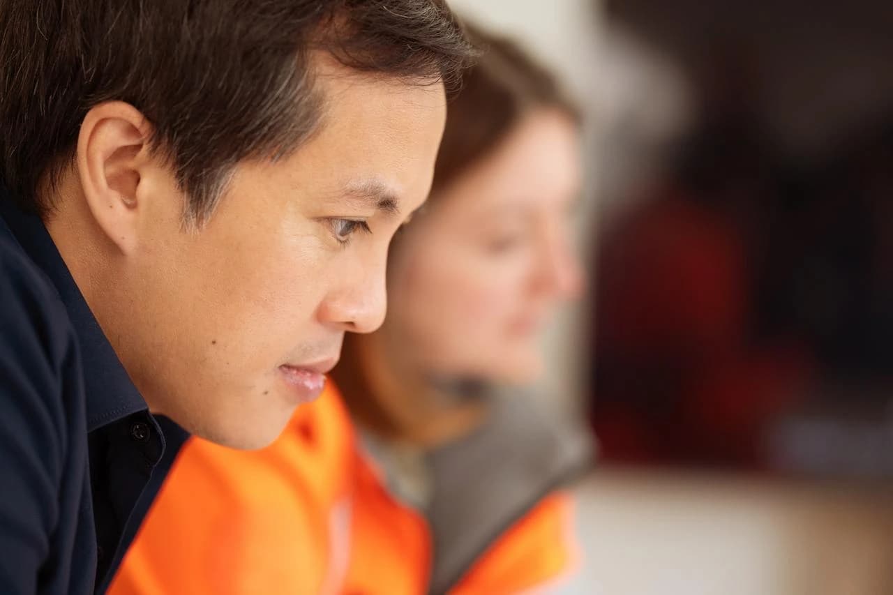 Gros plan sur le profil d'un homme concentré, portant une chemise bleue et un gilet orange, avec une femme floue en arrière-plan.