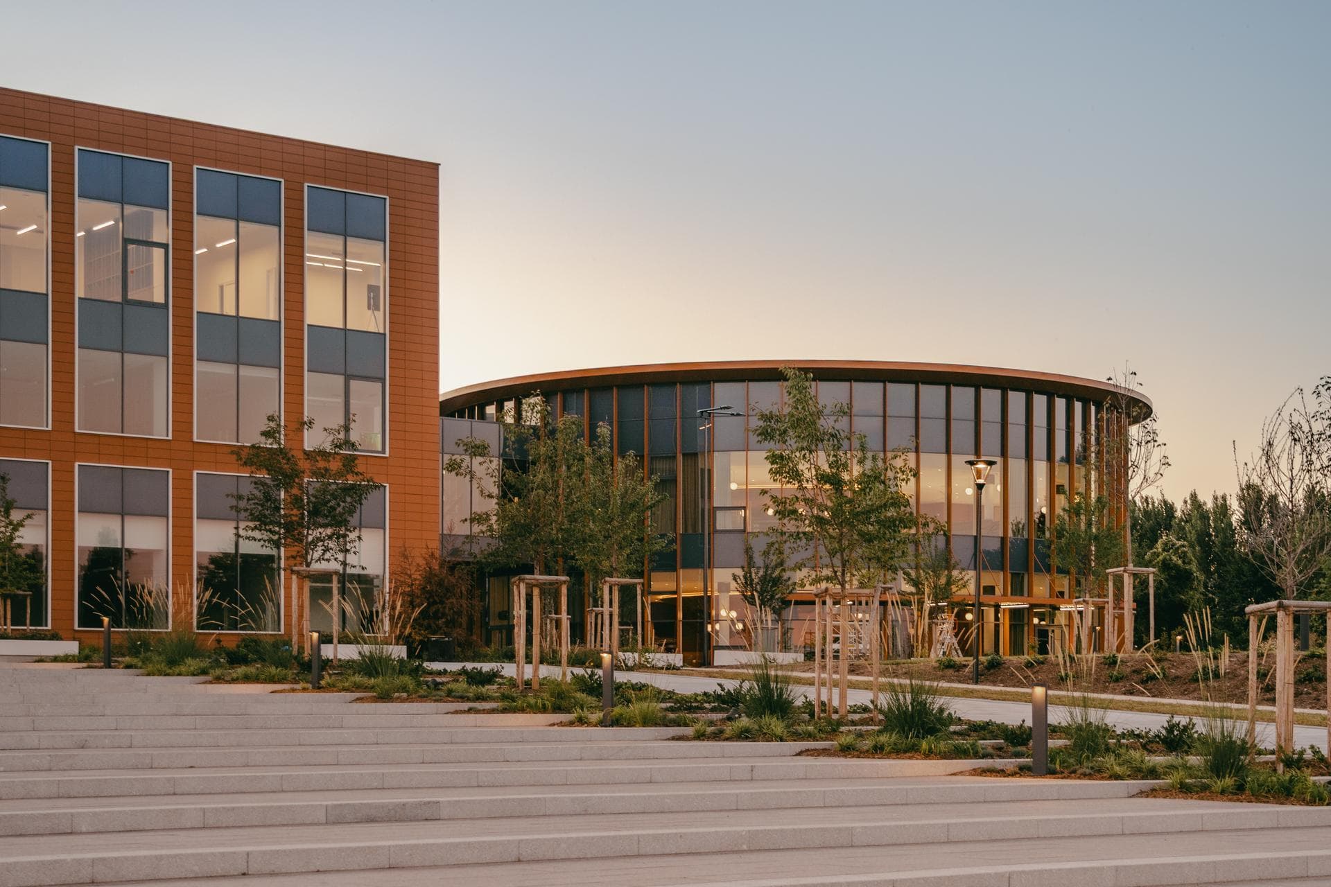 Une vue extérieure au crépuscule de deux bâtiments modernes, un rectangulaire avec des panneaux couleur terre cuite et un rond vitré, éclairés par l'intérieur et entourés d'aménagements paysagers avec un grand escalier en pierre au premier plan.
