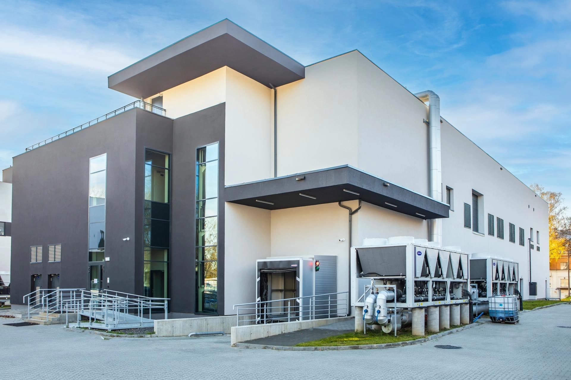Bâtiment moderne aux façades gris foncé et blanc cassé, doté de grandes fenêtres, d'une rampe d'accès, d'un quai de chargement et de systèmes de climatisation extérieurs, sous un ciel bleu lumineux.