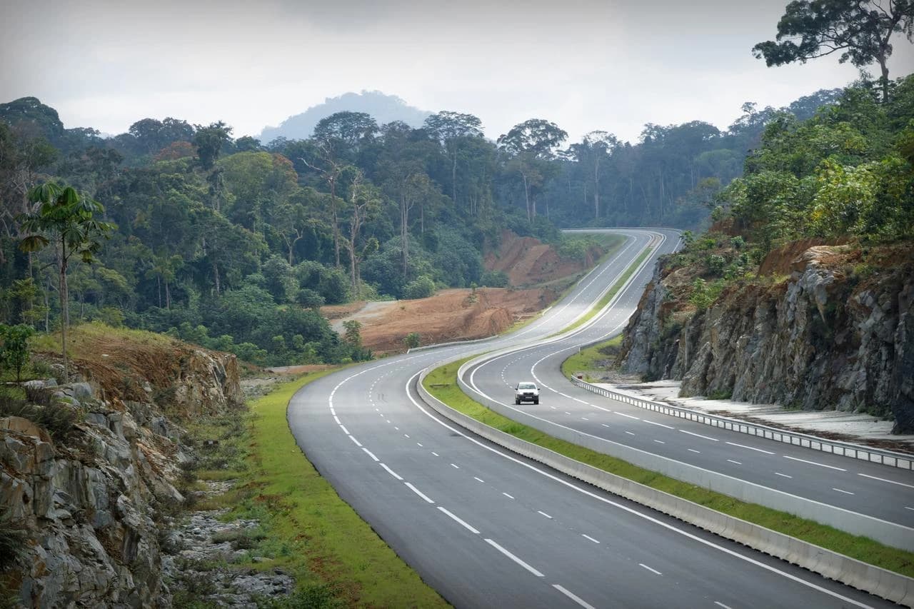 Une autoroute moderne et sinueuse à plusieurs voies traverse une dense forêt tropicale verdoyante sous un ciel gris, bordée par des falaises rocheuses et des talus de terre, avec une voiture blanche roulant au loin.