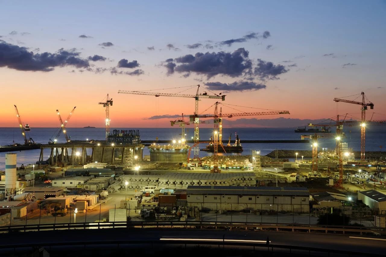 Vaste chantier de construction industrielle côtière au crépuscule, avec de nombreuses grues illuminées surplombant des structures en développement, des bâtiments et la mer sous un ciel dégradé d'orange et de bleu.