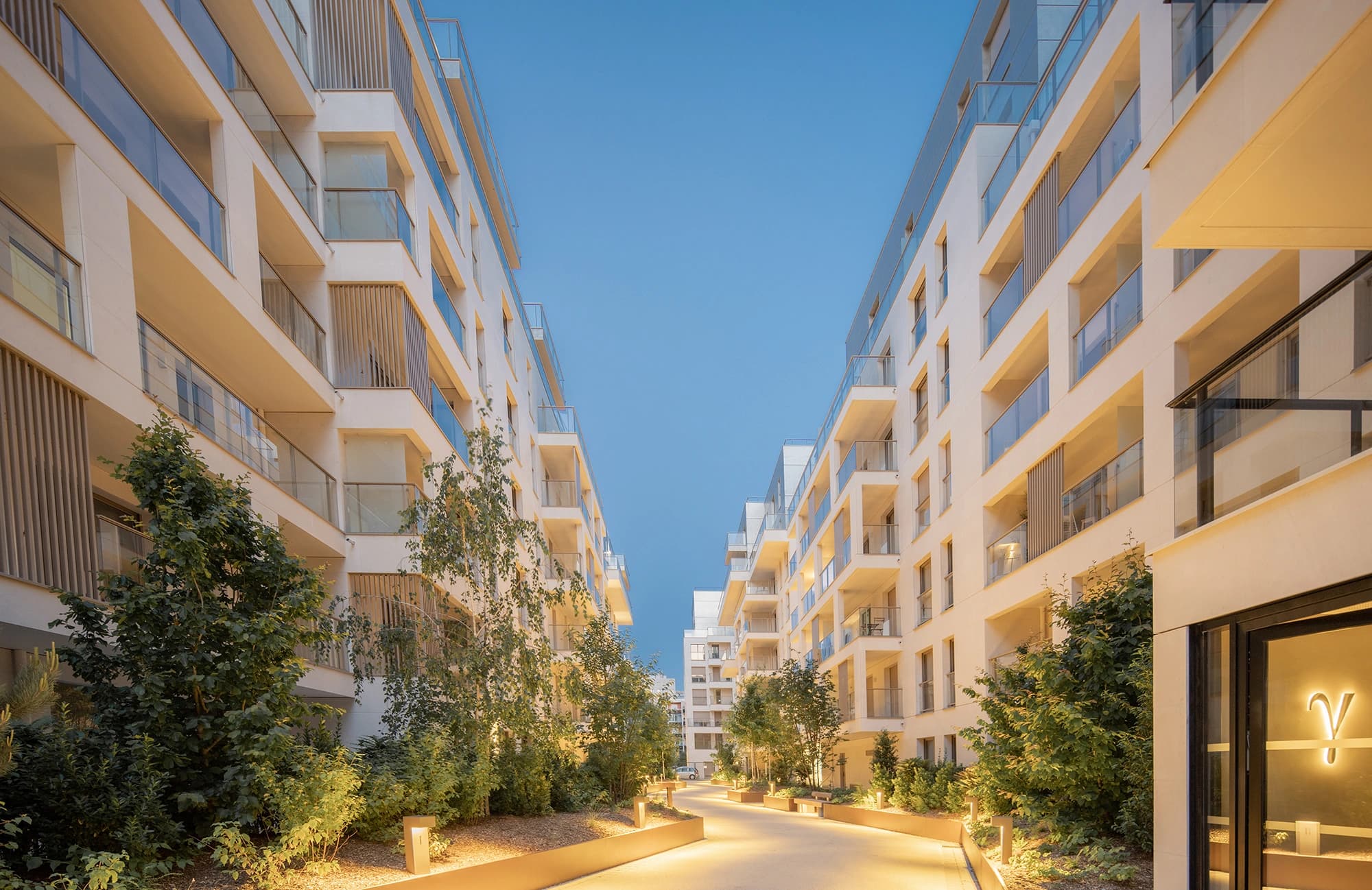 Un passage piéton moderne bordé de verdure et d'éclairages au sol chaleureux, flanqué de deux immeubles résidentiels blancs à balcons en verre sous un ciel bleu clair au crépuscule.