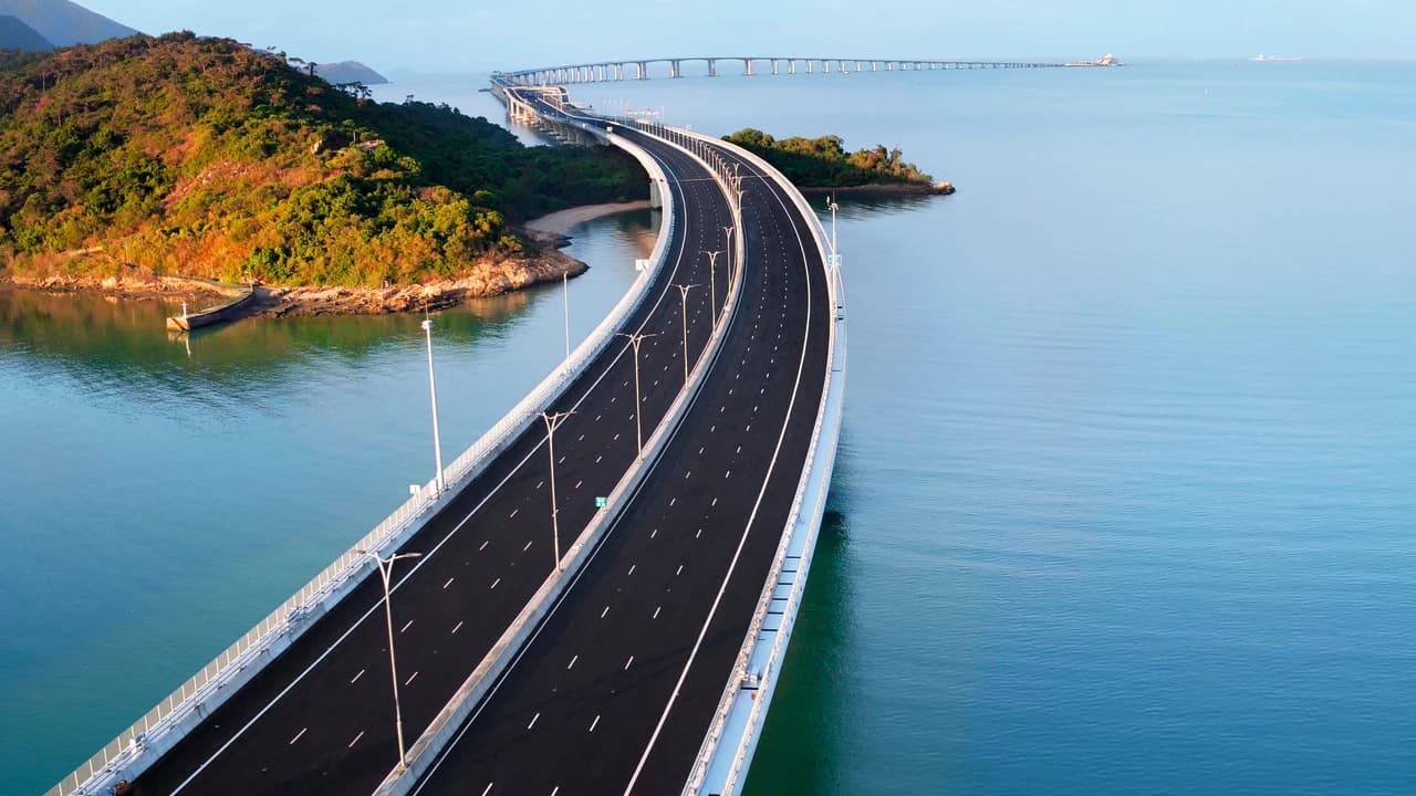 Un long pont autoroutier sinueux, bordé de lampadaires, traverse une étendue d'eau bleue et calme, reliant des collines verdoyantes et une île, puis s'étire vers l'horizon.