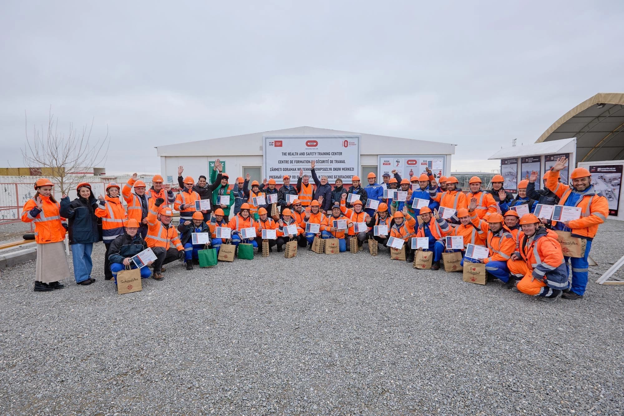 Un grand groupe de personnes, majoritairement des hommes, portant des vestes de sécurité et des casques de protection orange vif, sourient et agitent la main tout en tenant des certificats et des sacs cadeaux. Ils sont regroupés sur un sol de gravier devant un bâtiment blanc identifié comme un "Centre de formation à la santé et à la sécurité".