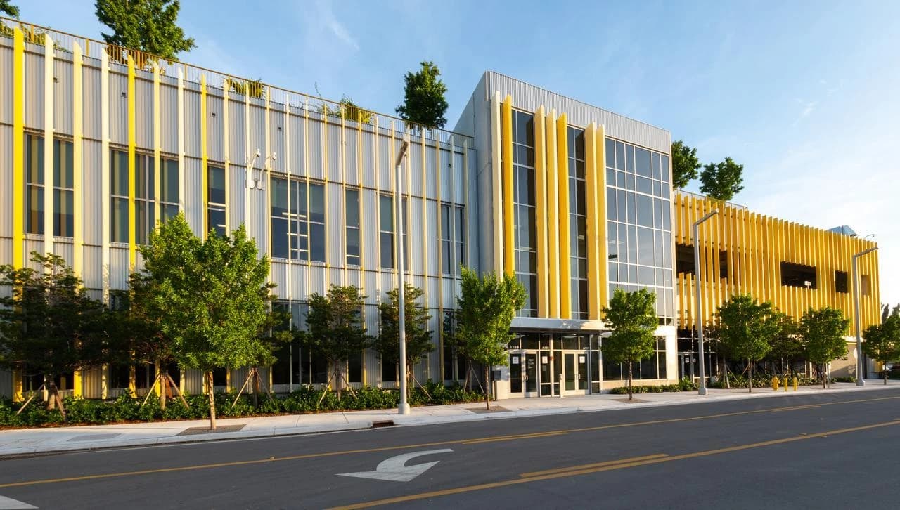 Modern building with a grey facade and bright yellow vertical accents, lined with trees and a street.