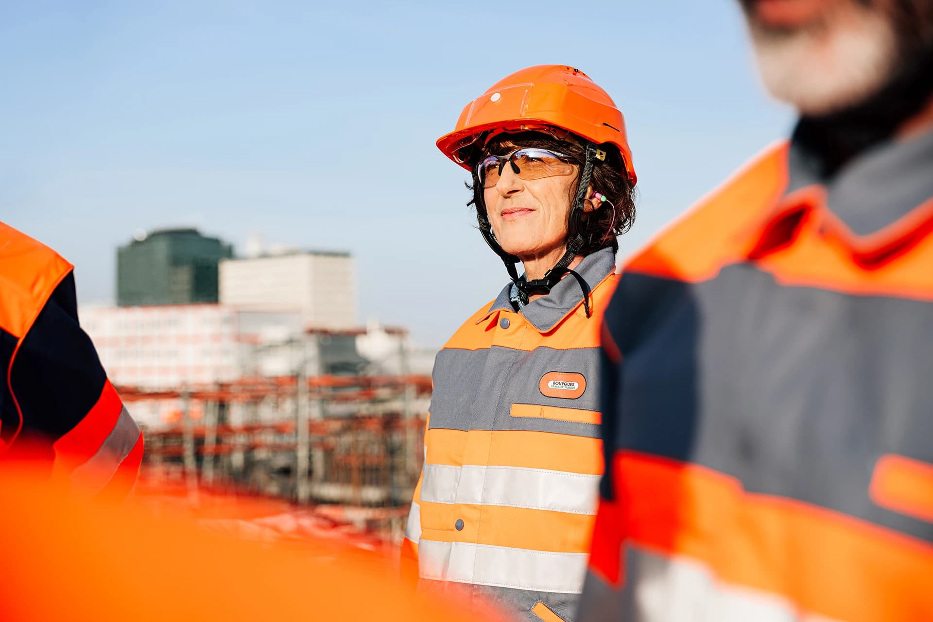 Femme souriante en casque de sécurité orange, lunettes de protection et veste de travail grise et orange à bandes réfléchissantes avec le logo Bouygues Construction, sur un chantier sous un ciel bleu clair.