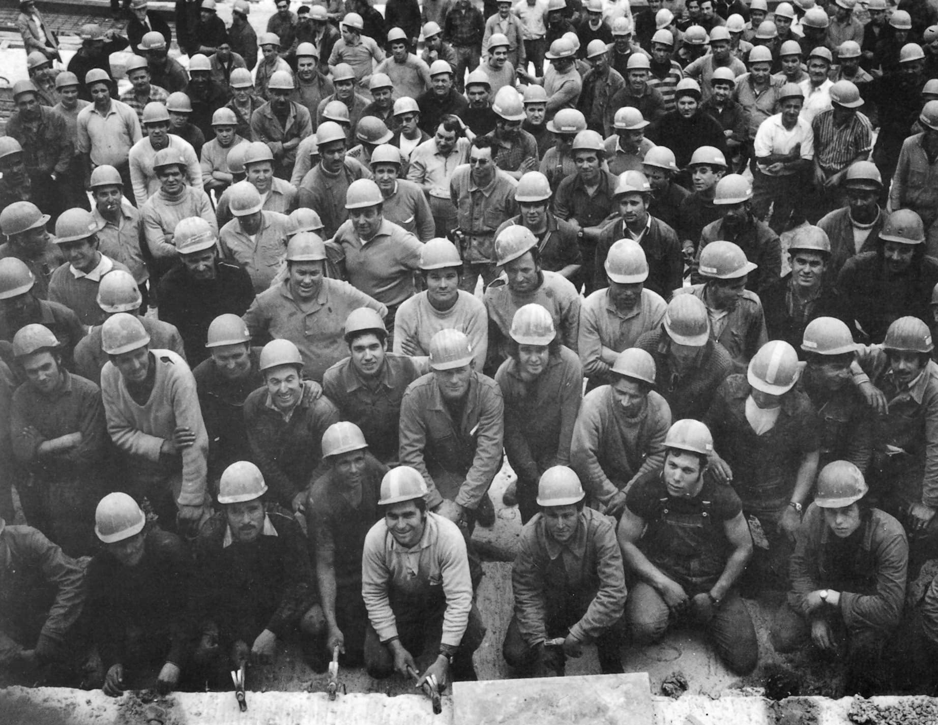Une photographie en niveaux de gris montre un grand groupe d'ouvriers du bâtiment souriants, beaucoup portant des casques de sécurité, posant en tenue de travail avec certains debout et d'autres accroupis au premier plan.