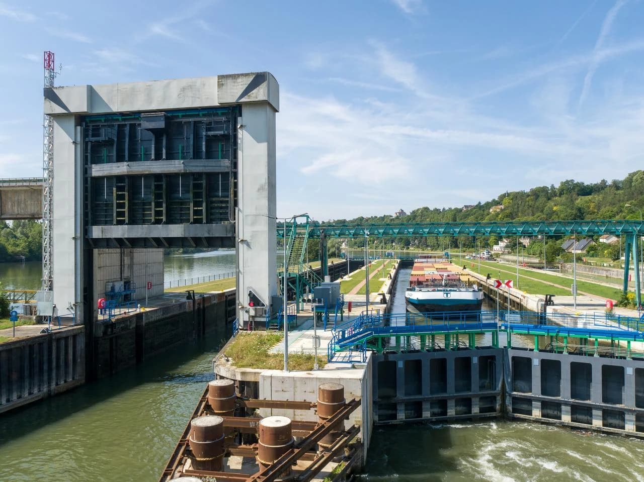 Grand ascenseur à bateaux en béton gris sur une rivière, avec un cargo amarré à l'intérieur sous un ciel bleu clair. Des collines verdoyantes et un aqueduc métallique vert s'étendent en arrière-plan.