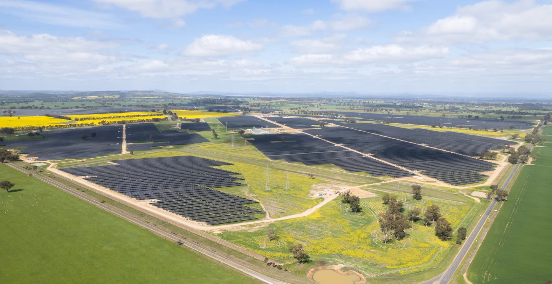 Vue aérienne d'une grande ferme solaire entourée de champs jaunes et verts sous un ciel clair et partiellement nuageux.