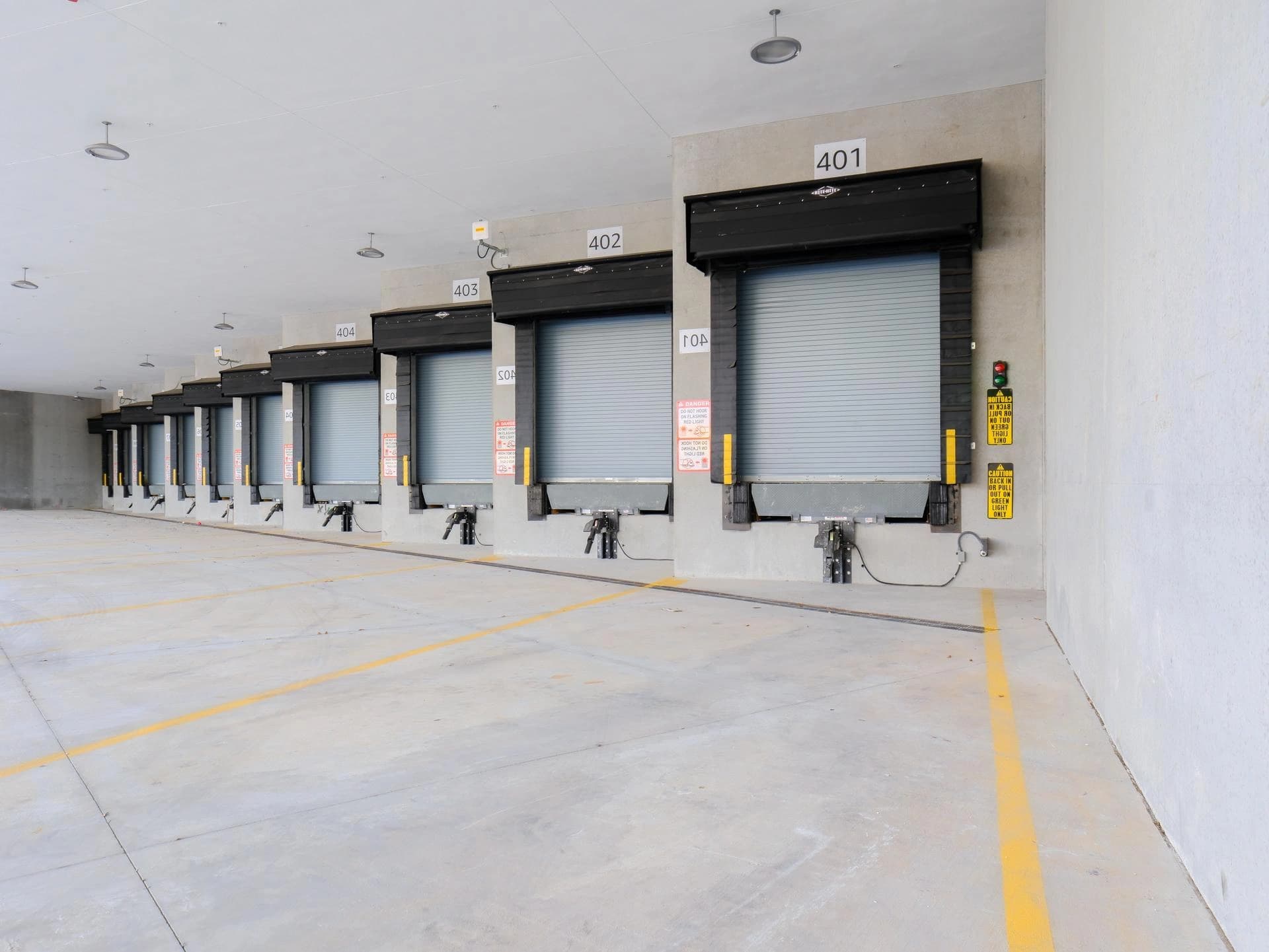 Une rangée de quais de chargement en béton avec des portes roulantes grises fermées, chacune numérotée, et des auvents noirs, sur un sol en béton gris clair marqué de lignes de sécurité jaunes.