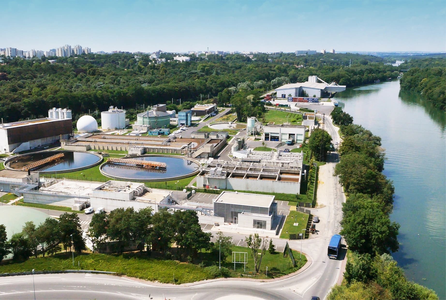 Vue aérienne d'une grande station d'épuration moderne, caractérisée par des bassins circulaires, des réservoirs blancs et divers bâtiments industriels. Elle est implantée au bord d'une large rivière et d'une forêt dense, avec une ville à l'horizon sous un ciel bleu.