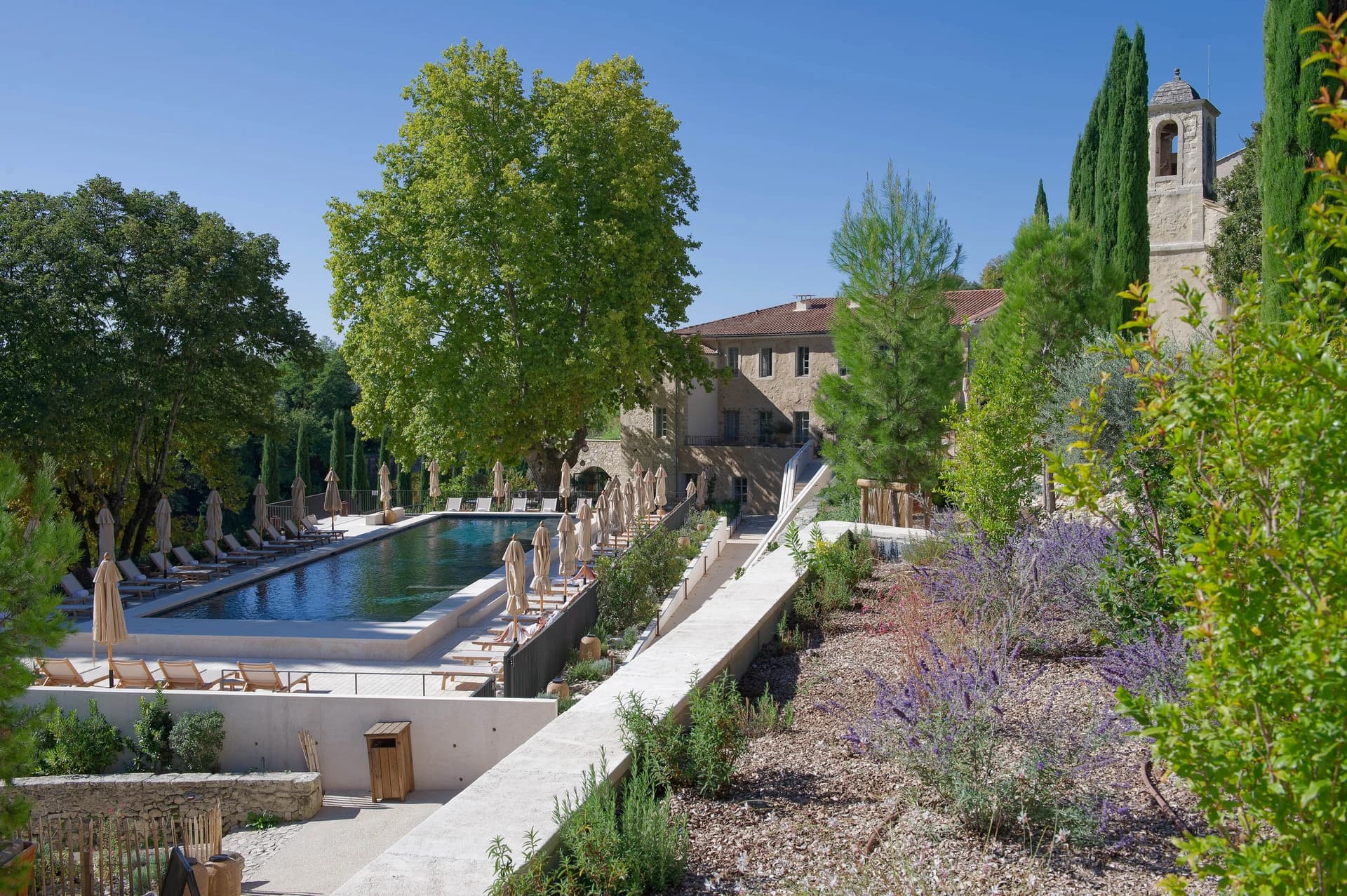 Vue panoramique d'une longue piscine bordée de transats et parasols beiges, située devant un bâtiment historique en pierre et un clocher. La scène est encadrée par de grands arbres verts luxuriants, des cyprès et des jardins en terrasses avec des plantes à fleurs violettes sous un ciel bleu clair.