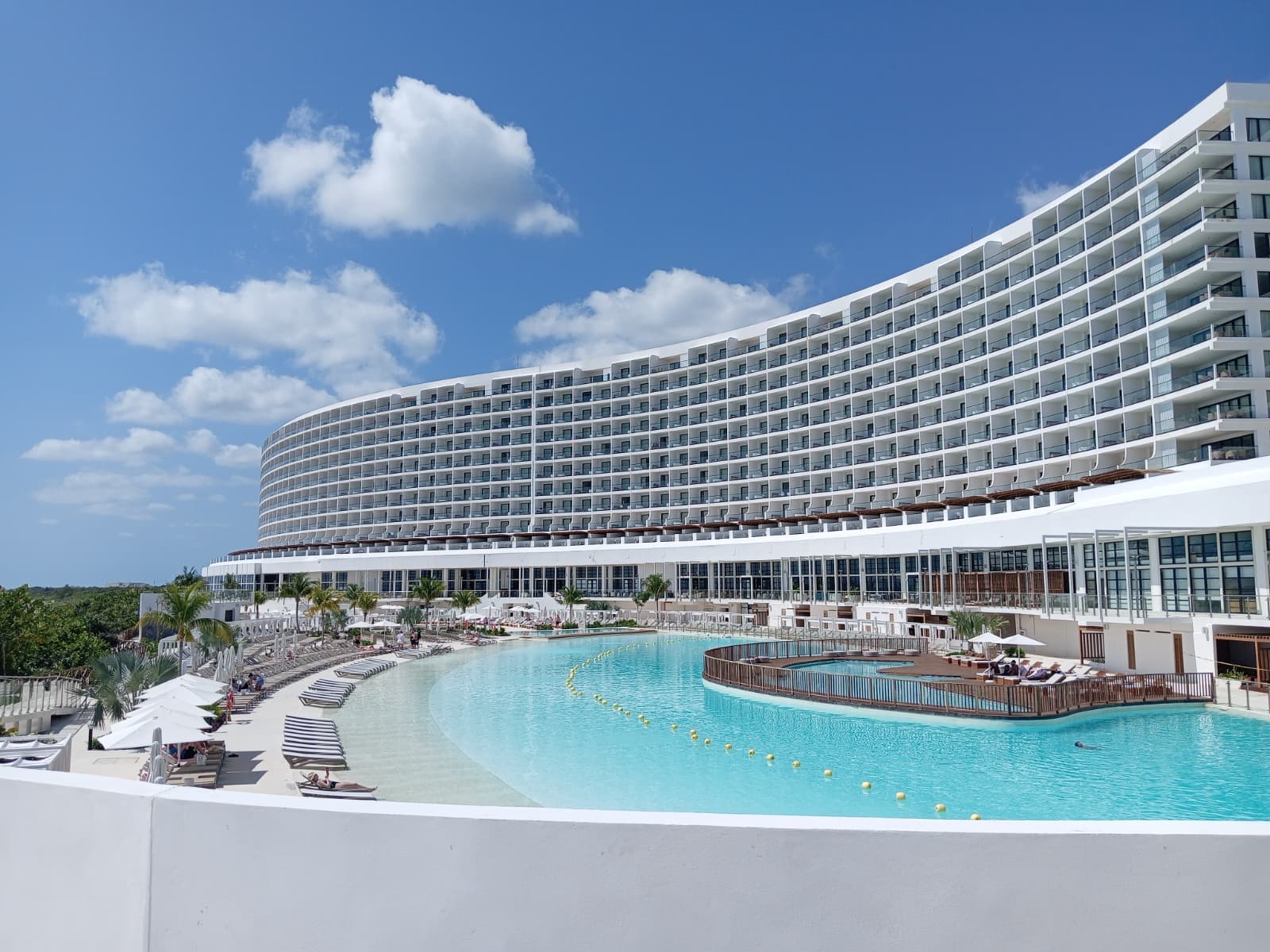 Un grand complexe hôtelier de luxe blanc et incurvé avec des centaines de balcons domine une immense piscine lagon bleu turquoise. Des chaises longues et des palmiers bordent le plan d'eau, sous un ciel bleu parsemé de nuages blancs, évoquant une destination de vacances ensoleillée.