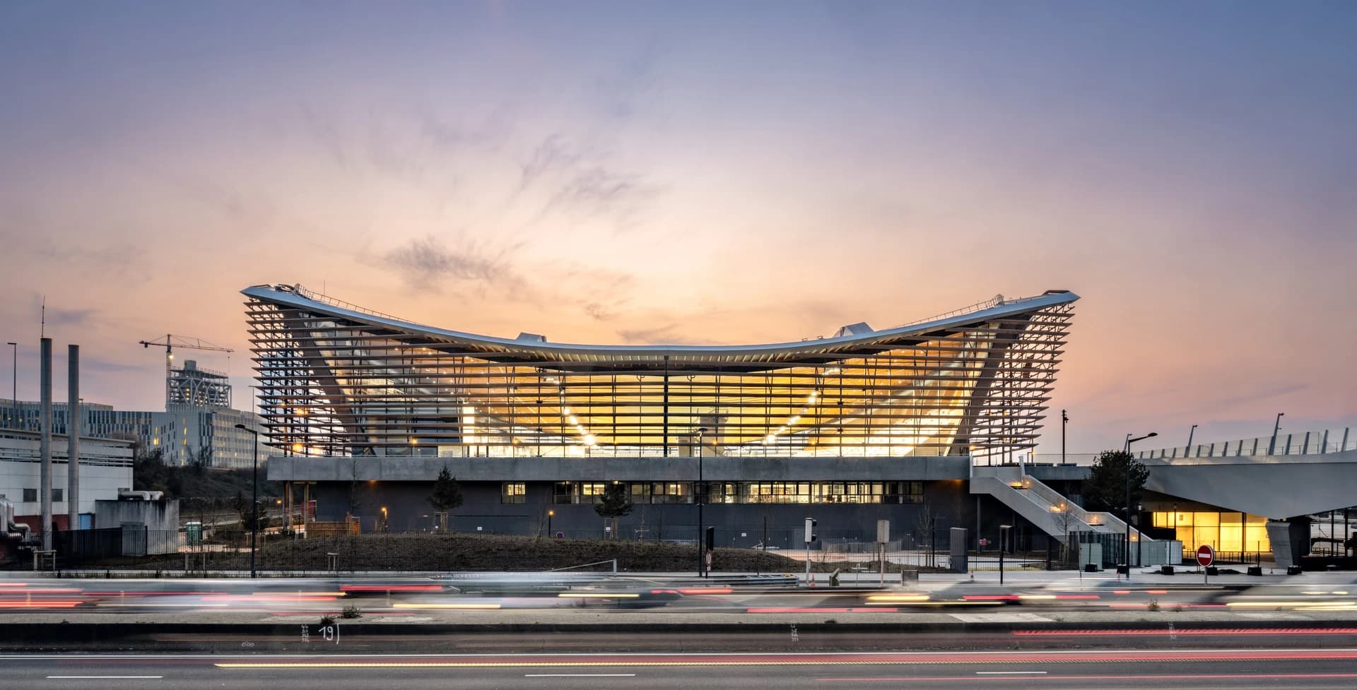 Un bâtiment moderne incurvé avec une façade de verre illuminée d'un orange chaud, se détache sur un ciel de crépuscule dégradé du rose-orange au bleu. Au premier plan, des traînées lumineuses de véhicules animent une route, soulignant le mouvement urbain.