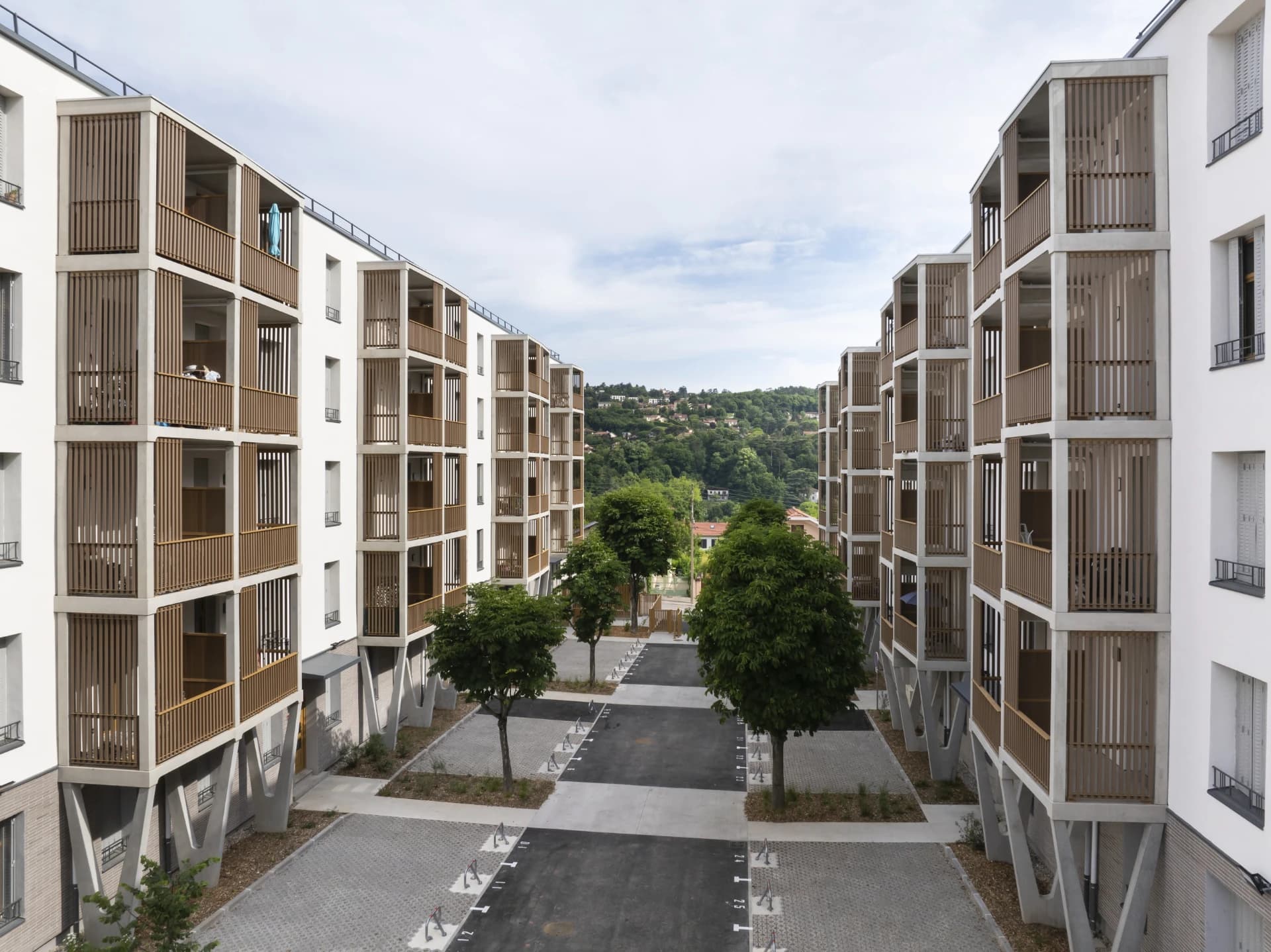 Des immeubles résidentiels modernes blancs aux balcons en béton avec écrans à lattes en bois encadrent une allée goudronnée bordée d'arbres verts. Au loin, une colline boisée parsemée de maisons se détache sous un ciel légèrement nuageux.