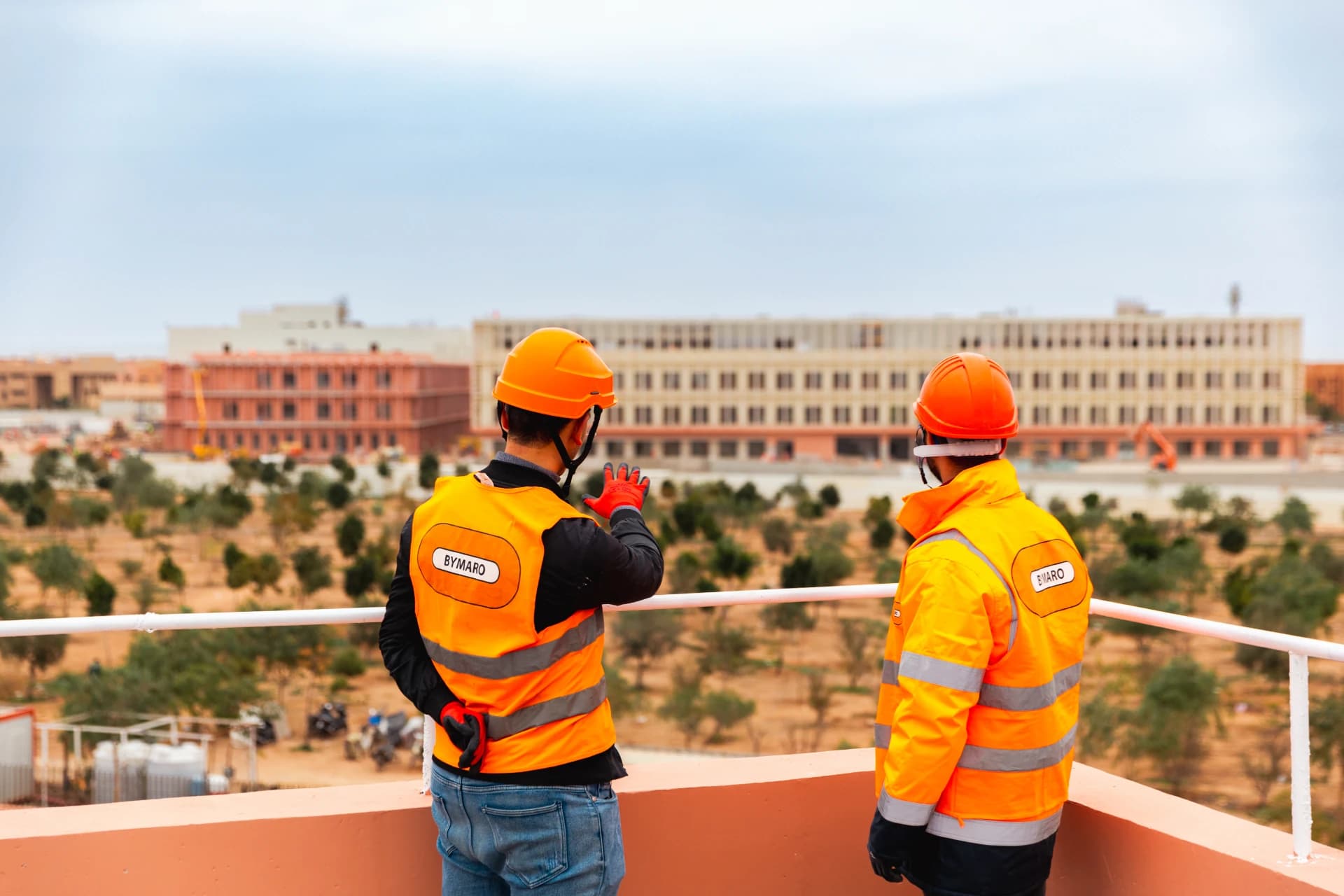 Deux hommes en casques et gilets de sécurité orange, dont l'un porte le logo BYMARO, observent un vaste chantier de construction depuis une terrasse, avec des bâtiments en cours d'édification et un paysage aride en arrière-plan.