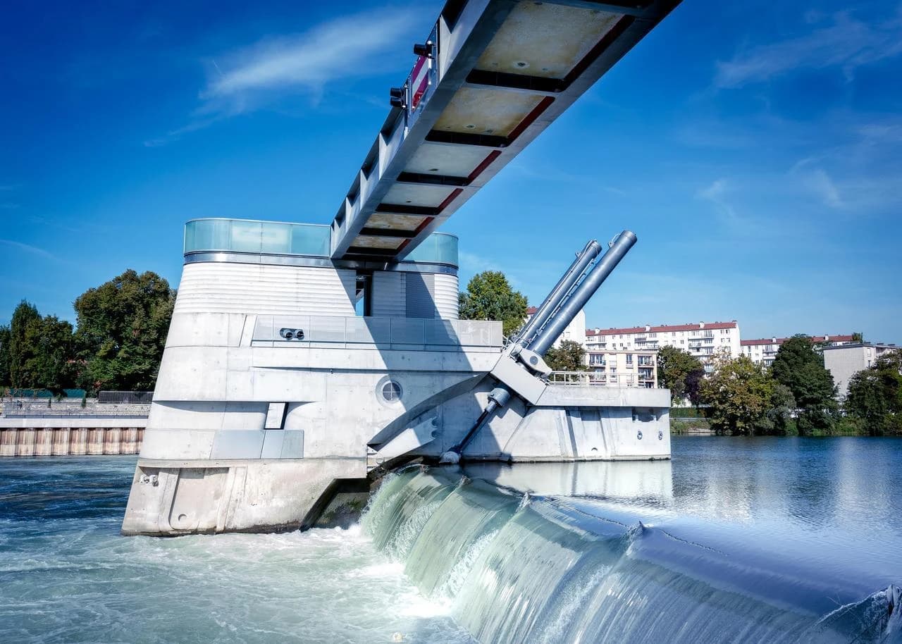 Un barrage moderne en béton gris clair avec des cylindres hydrauliques obliques crée une cascade d'eau écumeuse sous un ciel bleu clair. Une structure métallique inclinée le surplombe, avec des arbres verts et des bâtiments urbains en arrière-plan.