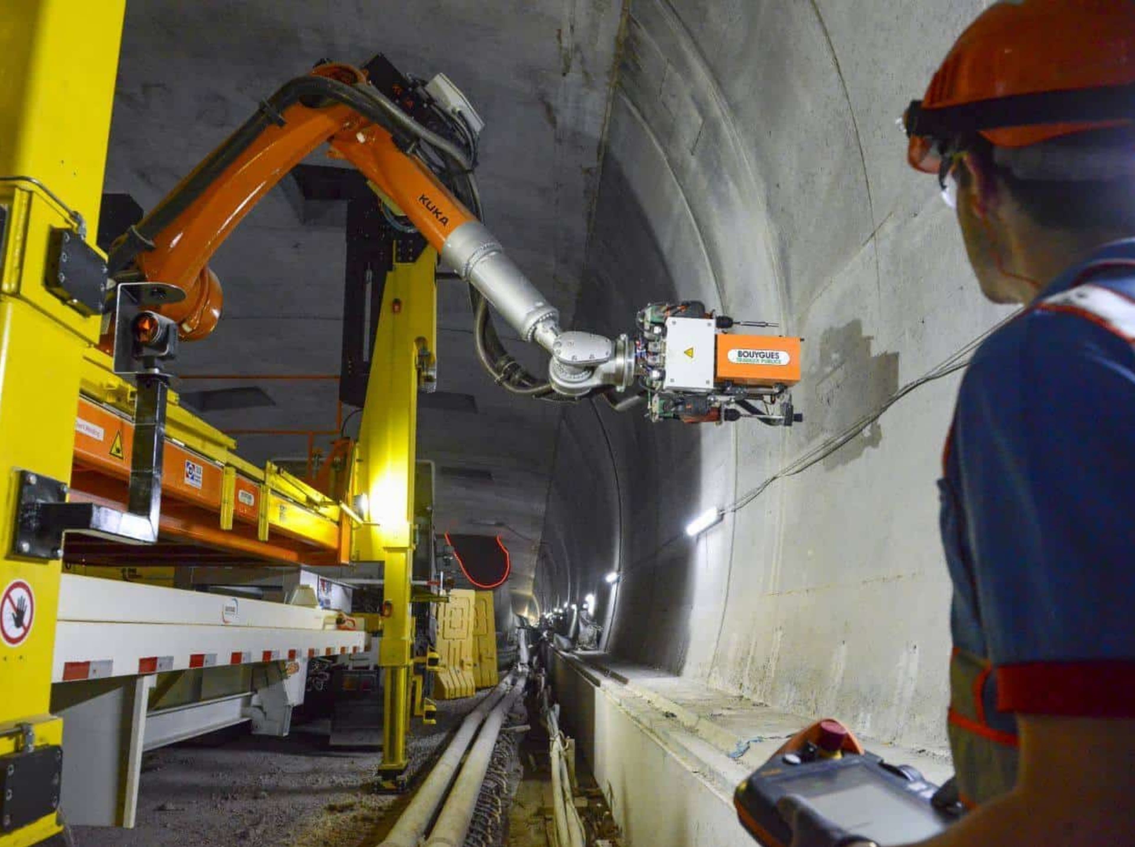 Un opérateur en casque orange contrôle un bras robotique KUKA orange et argent de Bouygues Travaux Publics, monté sur une structure jaune, effectuant des travaux le long de la paroi d'un tunnel en béton éclairé.
