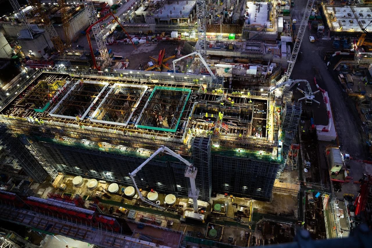 Vue aérienne nocturne d'un vaste chantier de construction animé, avec un grand bâtiment en édification entouré d'échafaudages, des grues et des ouvriers éclairés par des lumières intenses dans l'obscurité.