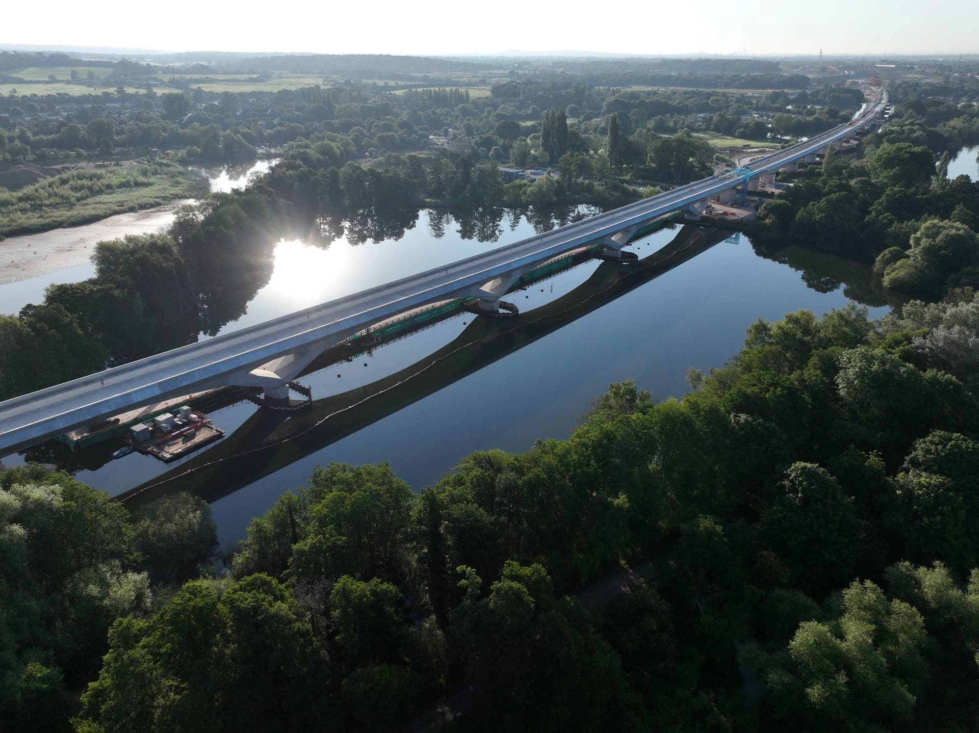 Vue aérienne d'un long pont ferroviaire moderne en construction traversant une rivière sombre aux reflets éclatants, entourée de forêts verdoyantes et de paysages ruraux. Des éléments de chantier sont visibles près des piles du pont.