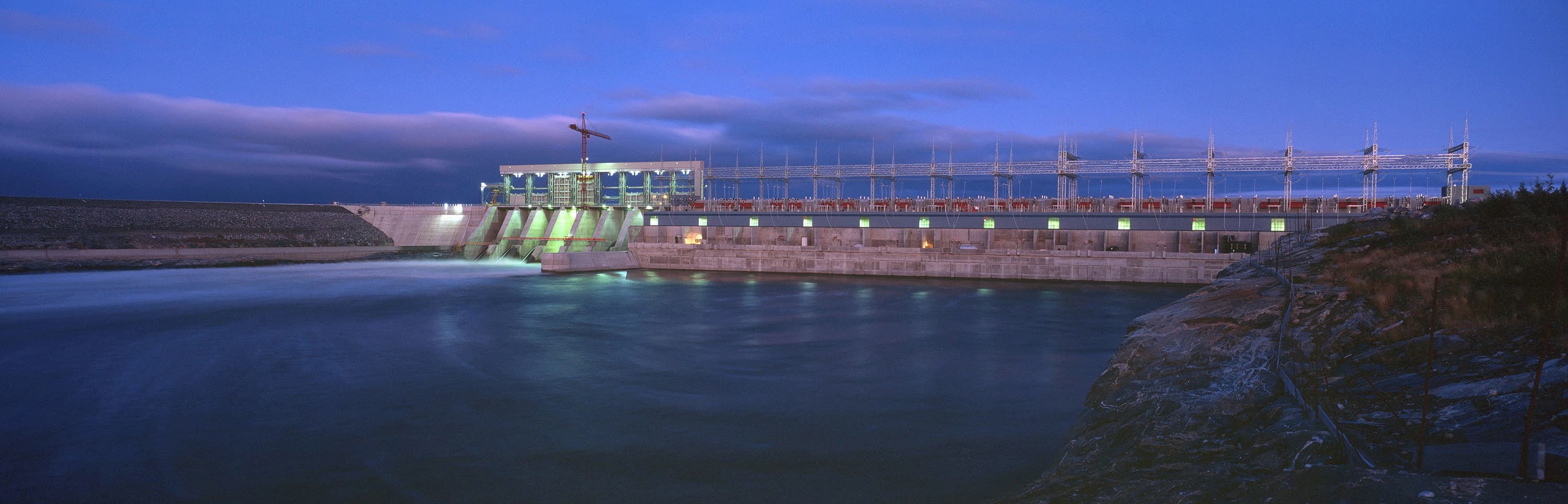 Vue nocturne d'un grand barrage hydroélectrique illuminé, avec de l'eau s'écoulant des vannes dans une rivière sous un ciel bleu foncé. Le complexe industriel comprend une centrale électrique avec des fenêtres lumineuses et une vaste infrastructure de lignes électriques en surface.