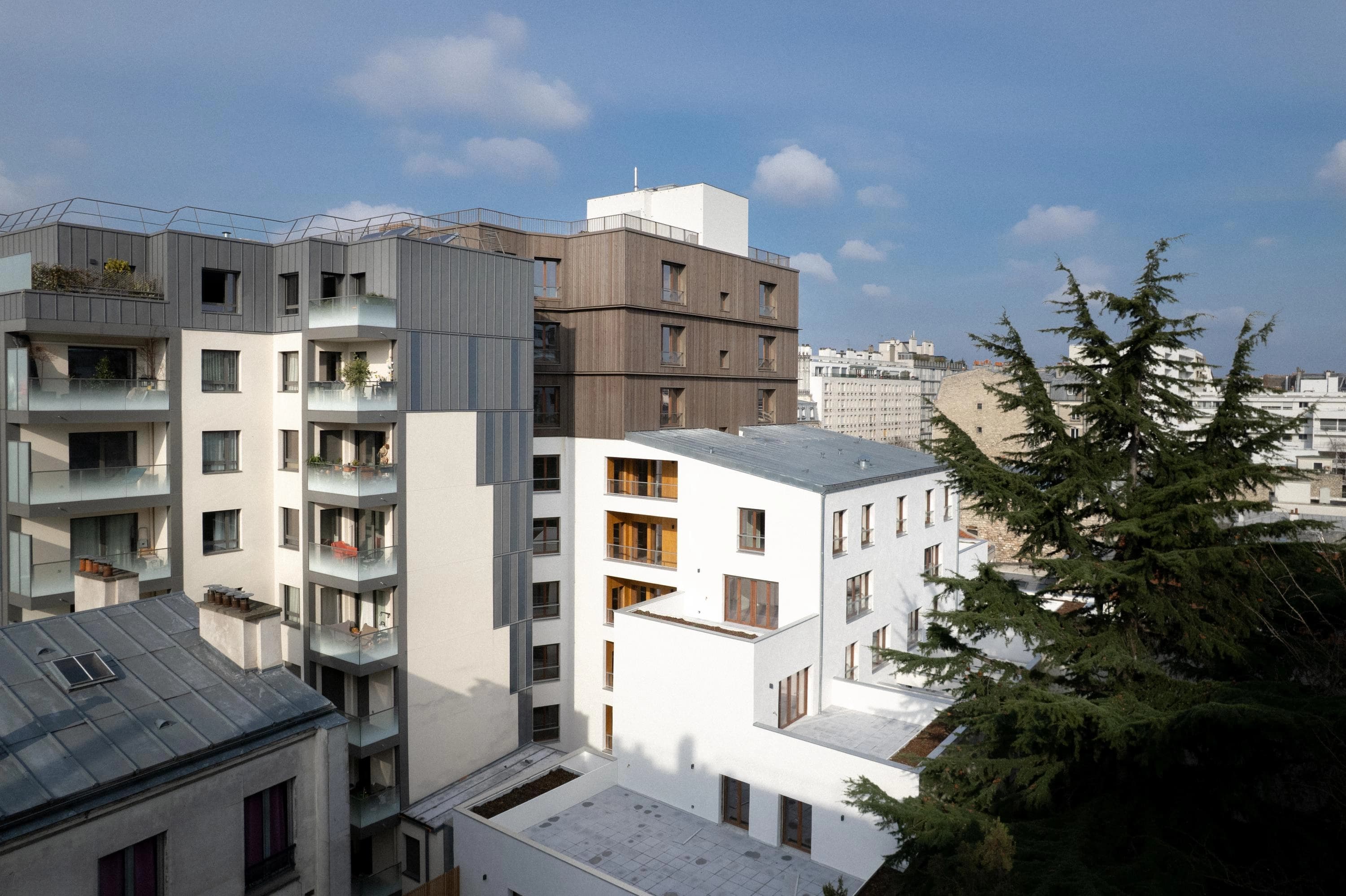 Ensemble de bâtiments résidentiels modernes aux façades gris métallique, beige, en bois brun et blanches, agrémenté de balcons en verre et encadré par des conifères verts sous un ciel bleu clair.