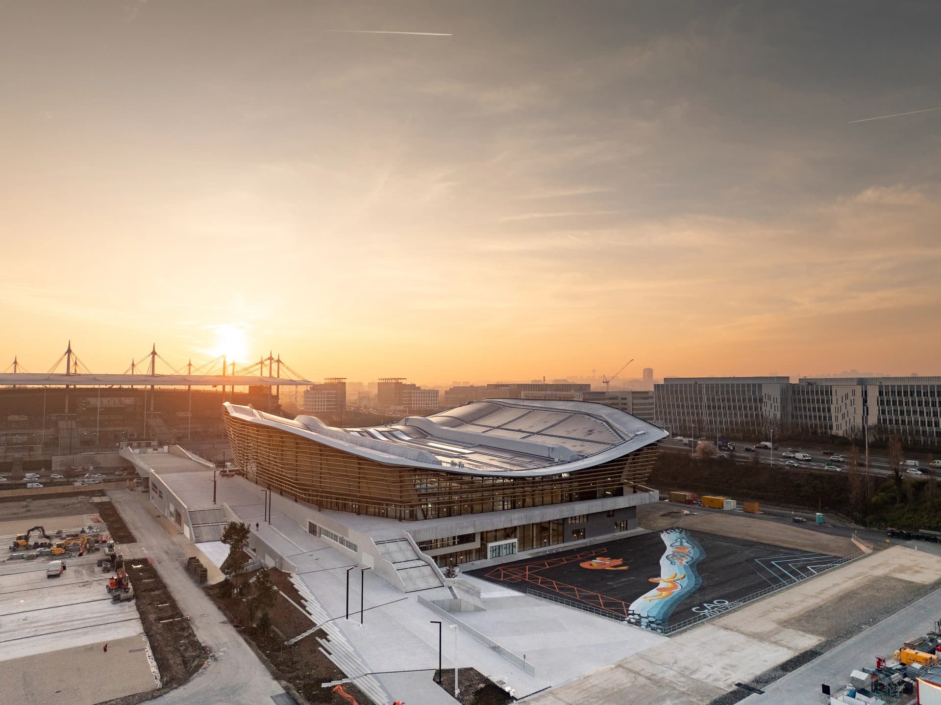 Une vue aérienne du Centre Aquatique Olympique moderne avec son toit incurvé, sa façade en verre et une grande place en béton, illuminée par le coucher de soleil doré, avec un pont à haubans et un paysage urbain en arrière-plan.