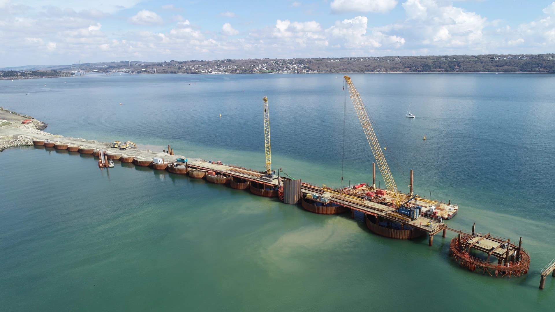 Vue aérienne d'un projet de construction de pont avec une longue estacade temporaire incurvée et des grues jaunes sur des coffrages circulaires s'avançant dans une étendue d'eau bleu-vert. Un pont suspendu lointain et une côte boisée parsemée de maisons sont visibles sous un ciel partiellement nuageux.