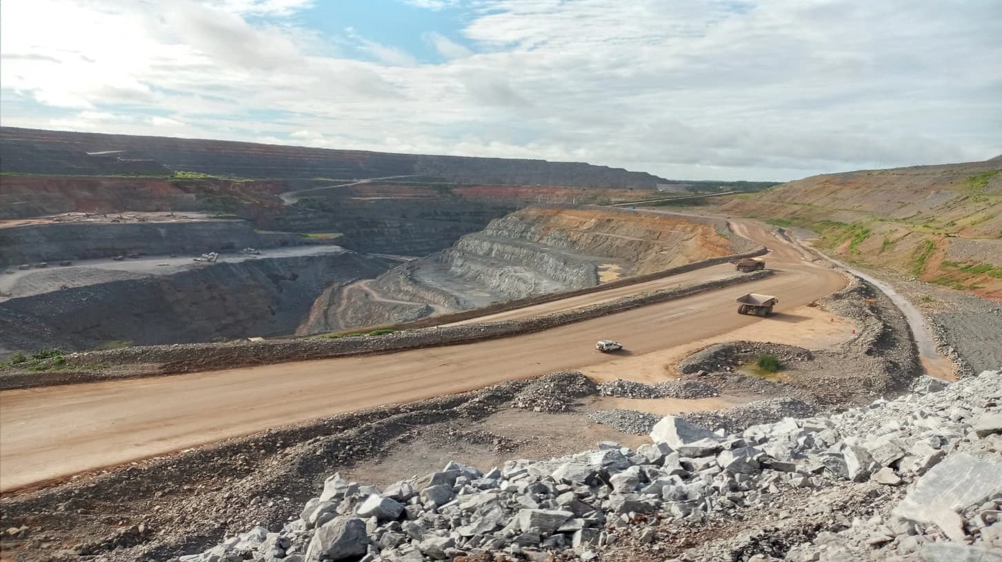 Vue aérienne d'une immense mine à ciel ouvert, montrant des terrasses de roche grise et rougeâtre, des routes en terre battue et plusieurs camions miniers sous un ciel nuageux.