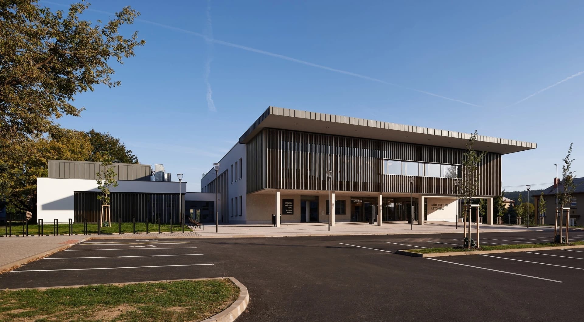 Un bâtiment moderne, abritant un centre culturel et une école d'art, présente une façade audacieuse avec des persiennes verticales sombres et des murs clairs, surmonté d'un large toit plat en porte-à-faux, avec un parking et des arbres au premier plan sous un ciel bleu clair.